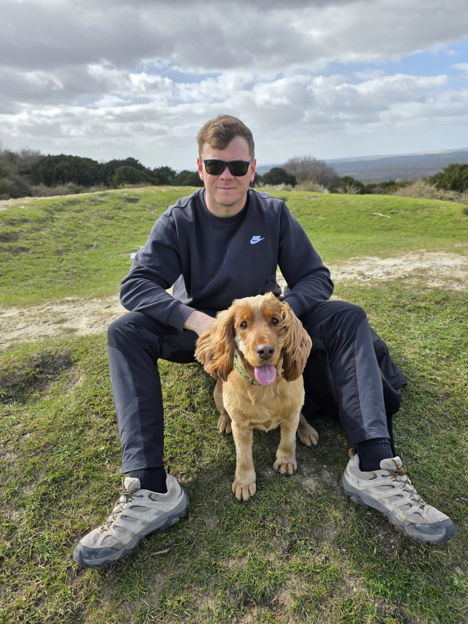 A man in sunglasses and a dark Nike sweatshirt sitting on grass with a cocker spaniel dog in a natural outdoor setting under a partly cloudy sky.