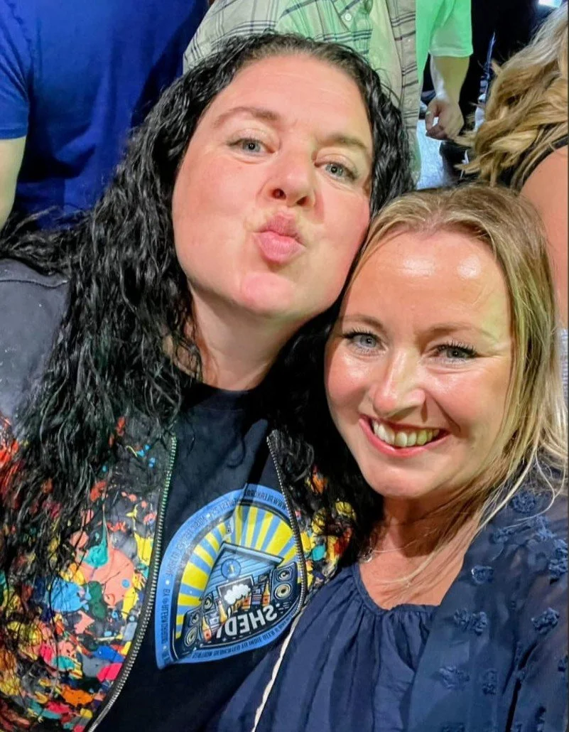 Two women smiling, taking a selfie together at a lively event with others in the background.