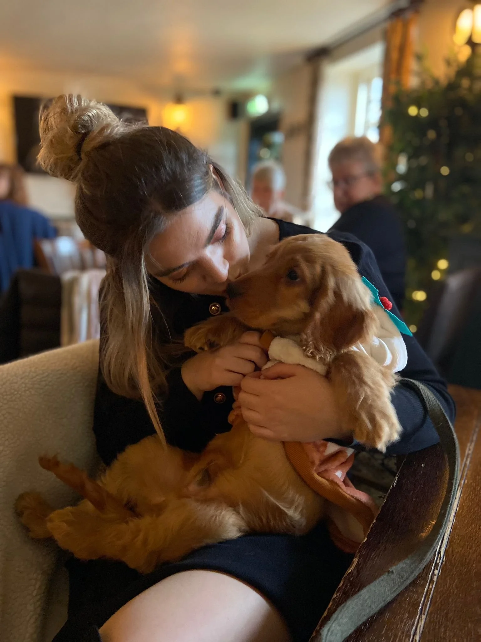 A woman with long hair in a bun is holding and kissing a cocker spaniel puppy in a cozy indoor setting, with several people and a Christmas tree in the background.