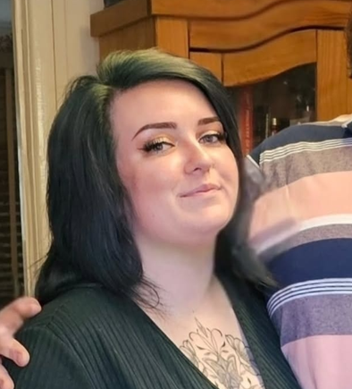A woman with dark green and black hair, wearing makeup and a black top, standing indoors near a wooden cabinet.