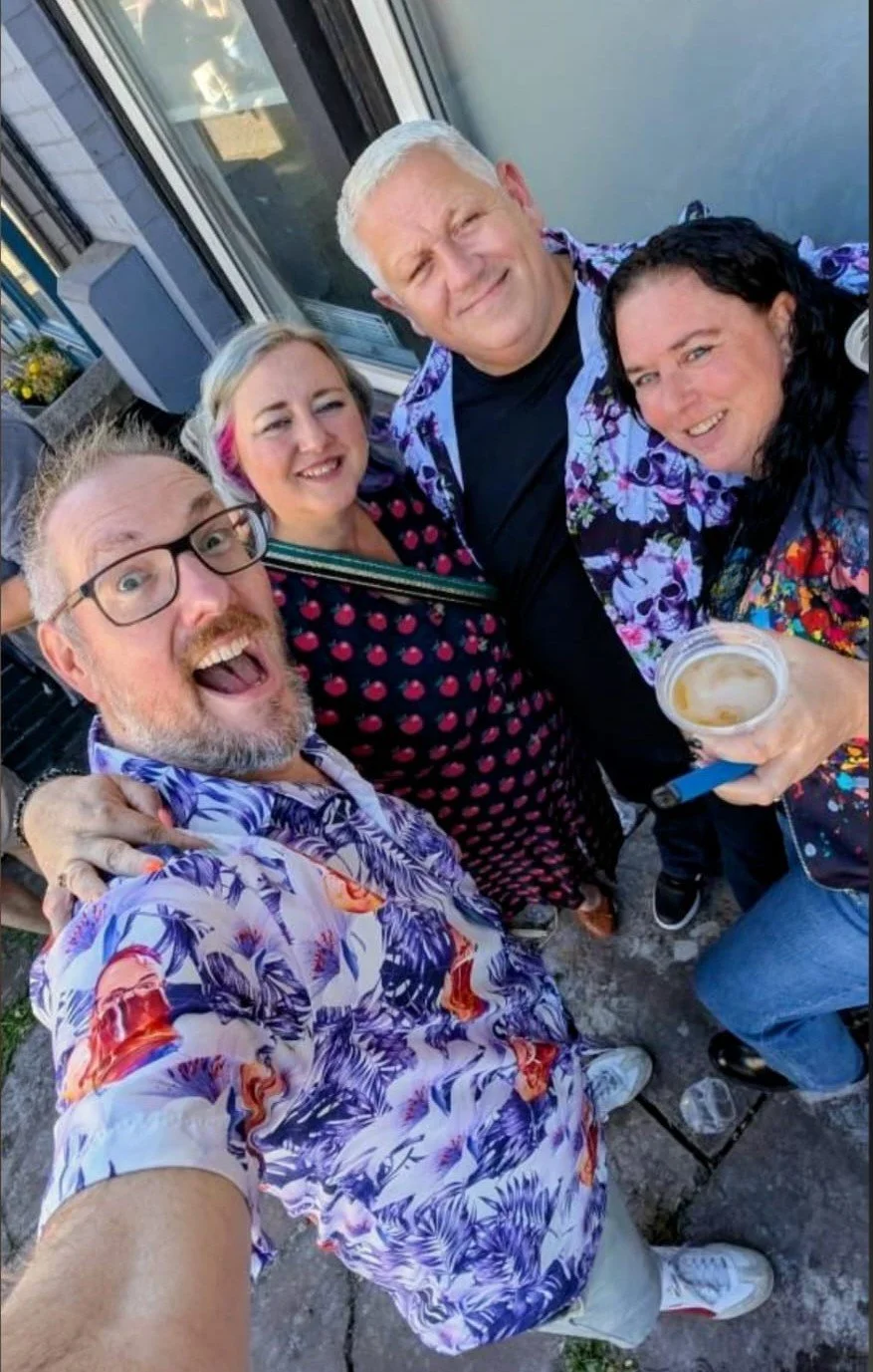 Group of five friends smiling and taking a selfie outdoors, dressed in colorful shirts with floral and fruit patterns, some holding a drink, standing on a stone patio near a building with a window.