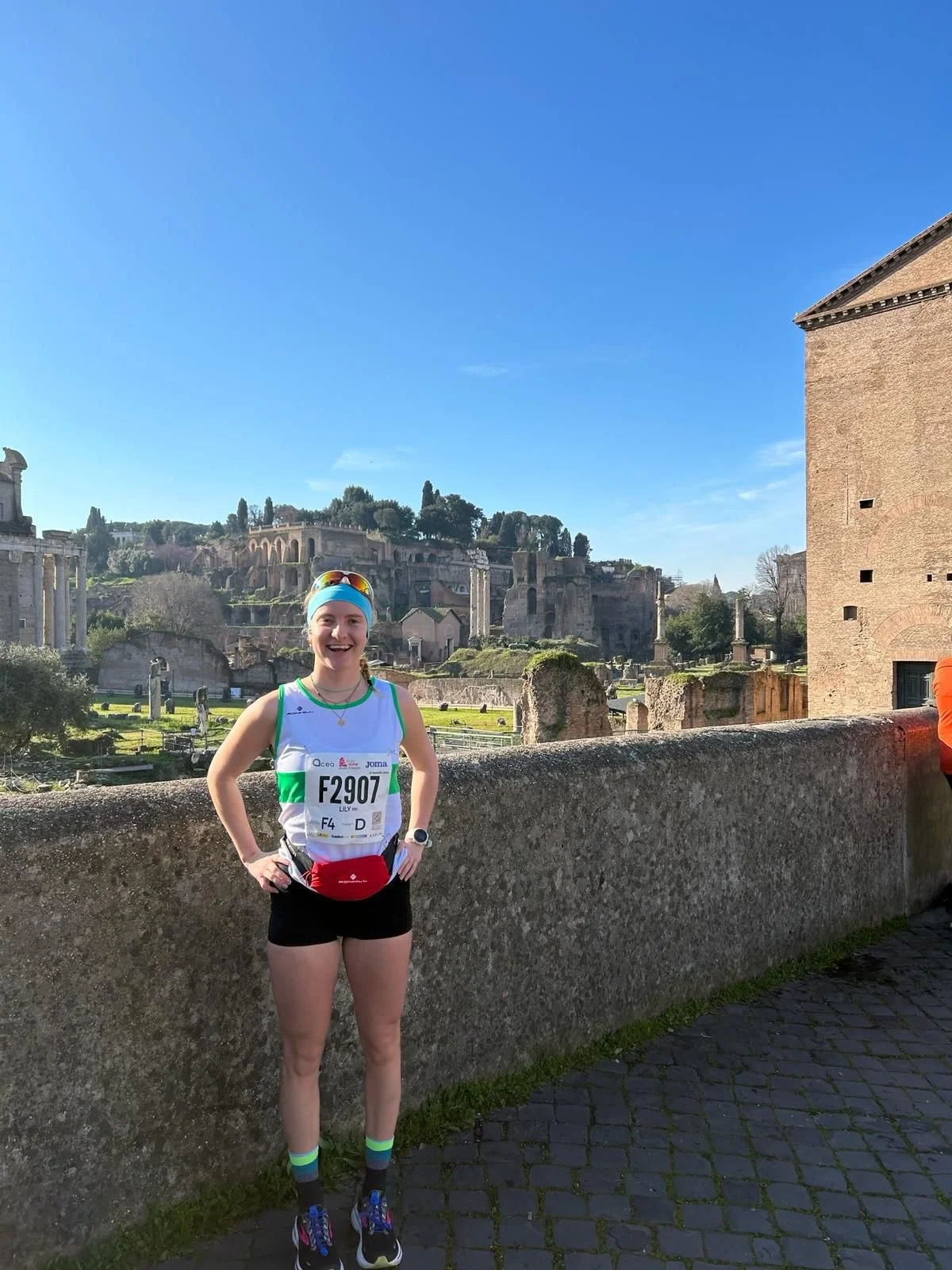 A woman in athletic attire standing outdoors on a stone pathway, smiling, with ancient ruins and a blue sky in the background.