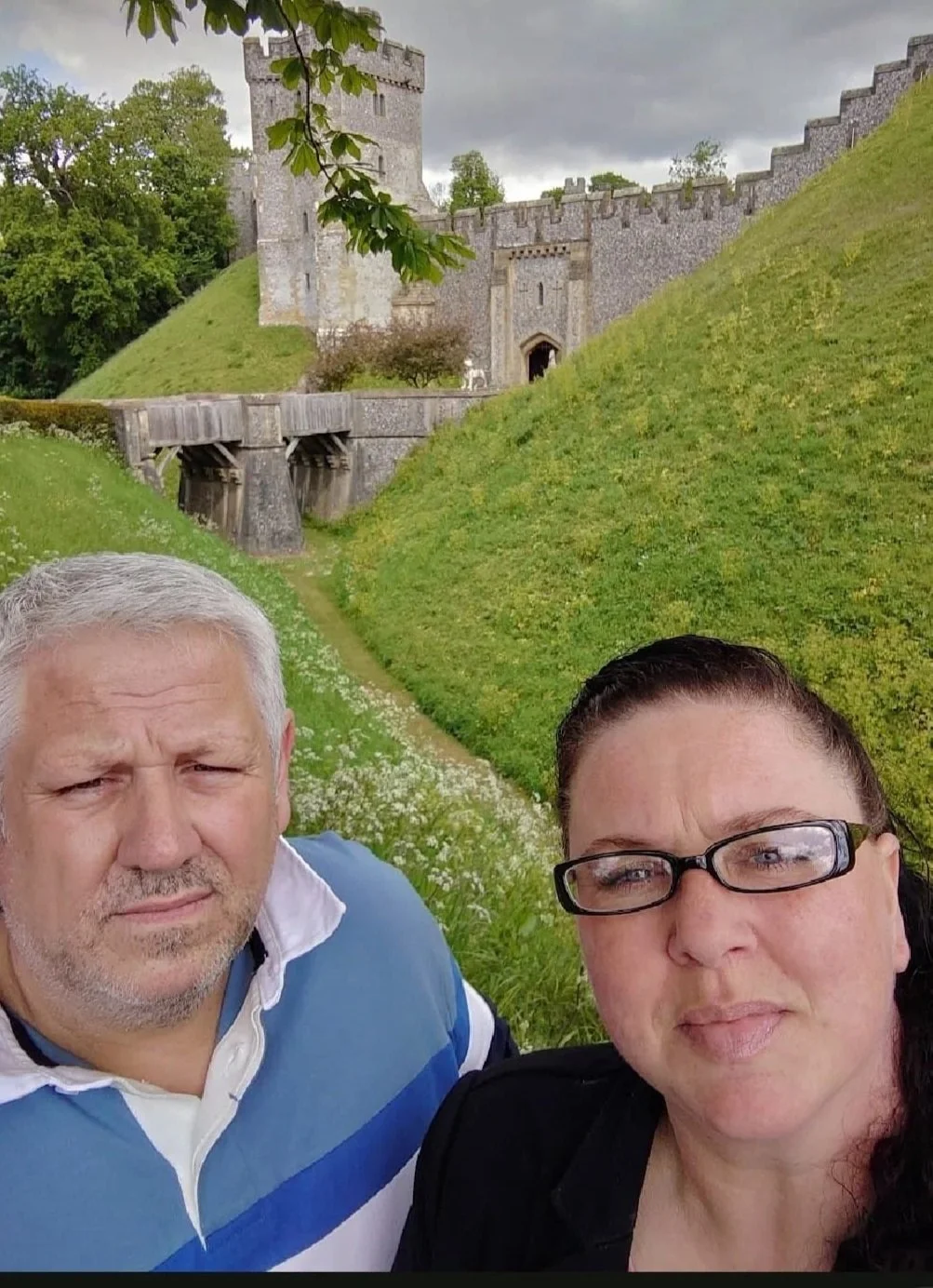A selfie of a man with short gray hair and a woman with dark hair and glasses, in front of a castle on a grassy hillside with cloudy sky.