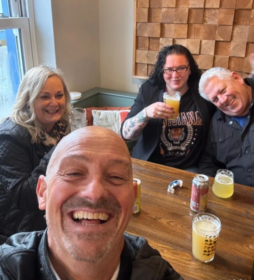 Four friends sitting at a wooden table, smiling and enjoying drinks in a cozy cafe or restaurant with a wooden wall and a window in the background.
