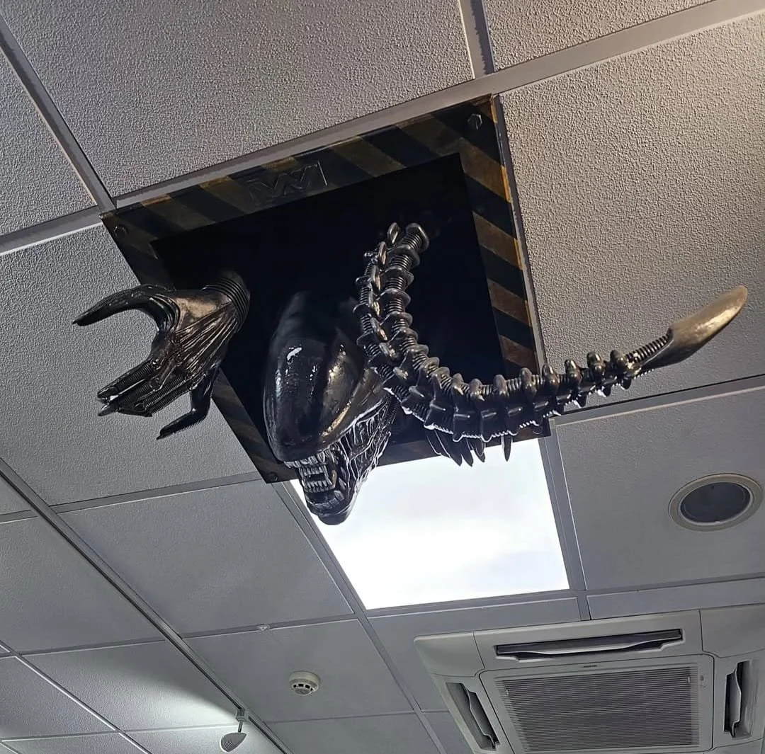 Alien creature sculpture emerging from ceiling vent, with a skull-like head, elongated limbs, and a tail.