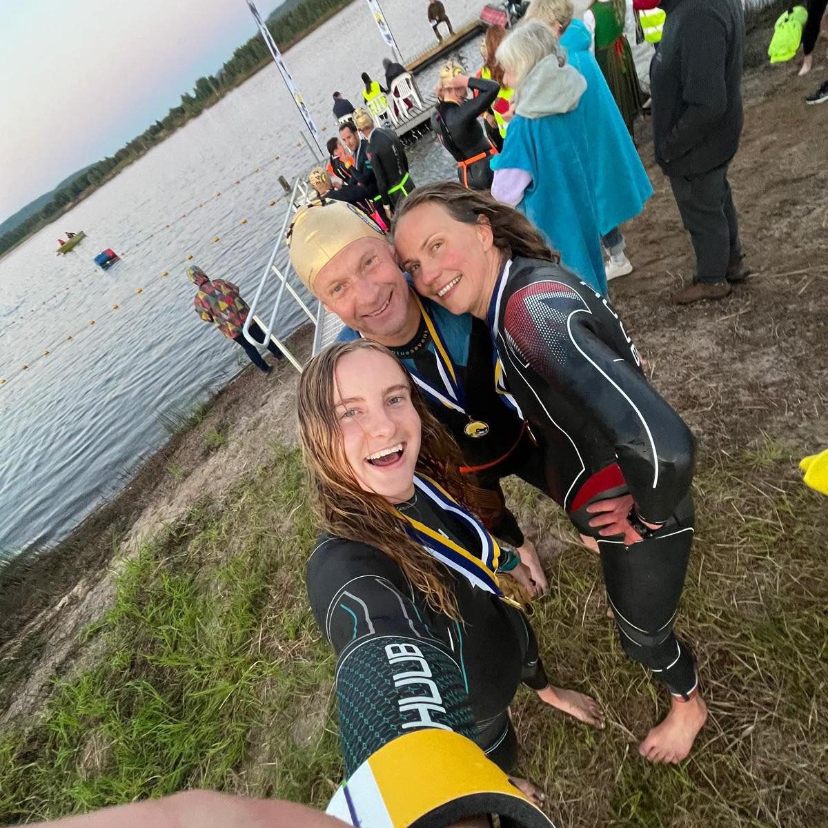 Three people taking a celebratory selfie on a beach after a water sports event, with a peaceful lake and a boat in the background, and other participants in racing gear nearby.