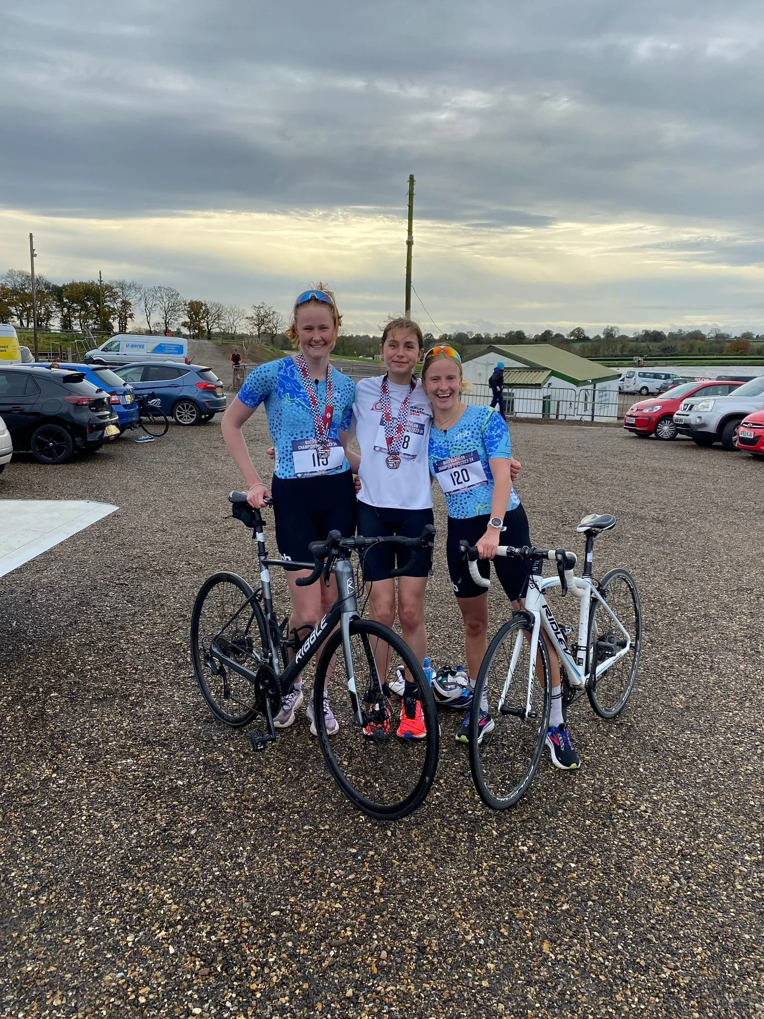 Three young women in athletic gear with medals around their necks, standing with bicycles in a parking lot after a race. All are smiling, and they are dressed in blue and white sportswear. Parking lot has cars and bikes, with cloudy sky overhead.