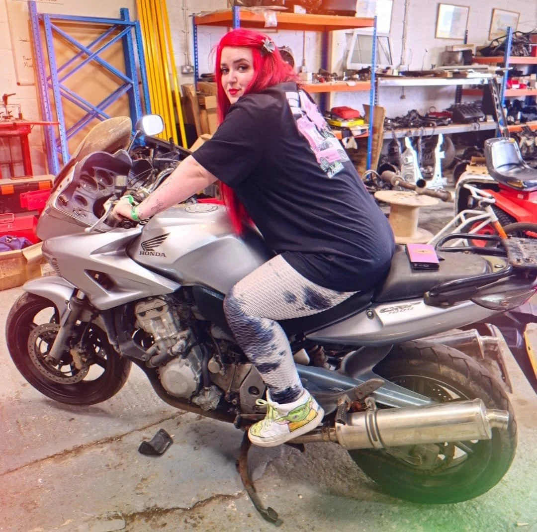 A woman with long red hair, wearing a black T-shirt and patterned leggings, sitting on a silver Honda motorcycle inside a workshop with shelves and tools.