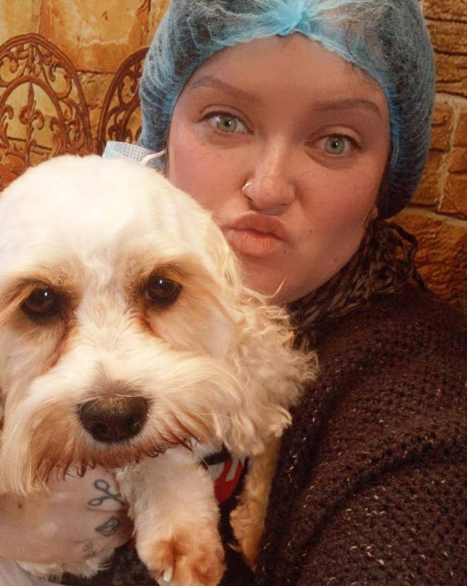 A woman with a blue hair cap, nose piercing, and light makeup taking a selfie with her dog in front of a brick wall. The dog has light-colored, curly fur and appears to be a small breed.