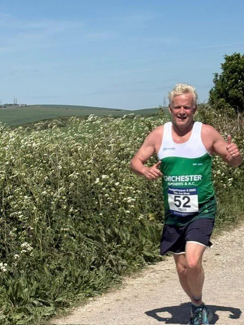 A man running outdoors on a gravel trail next to tall grass and wildflowers, smiling and giving a thumbs-up, wearing a green and white tank top with the number 52.