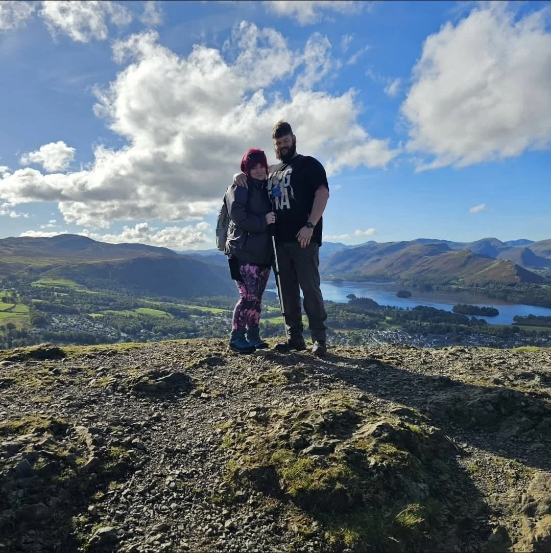 A couple standing on a rocky hilltop with scenic mountains in the Lake District , a lake, and a partly cloudy sky in the background.