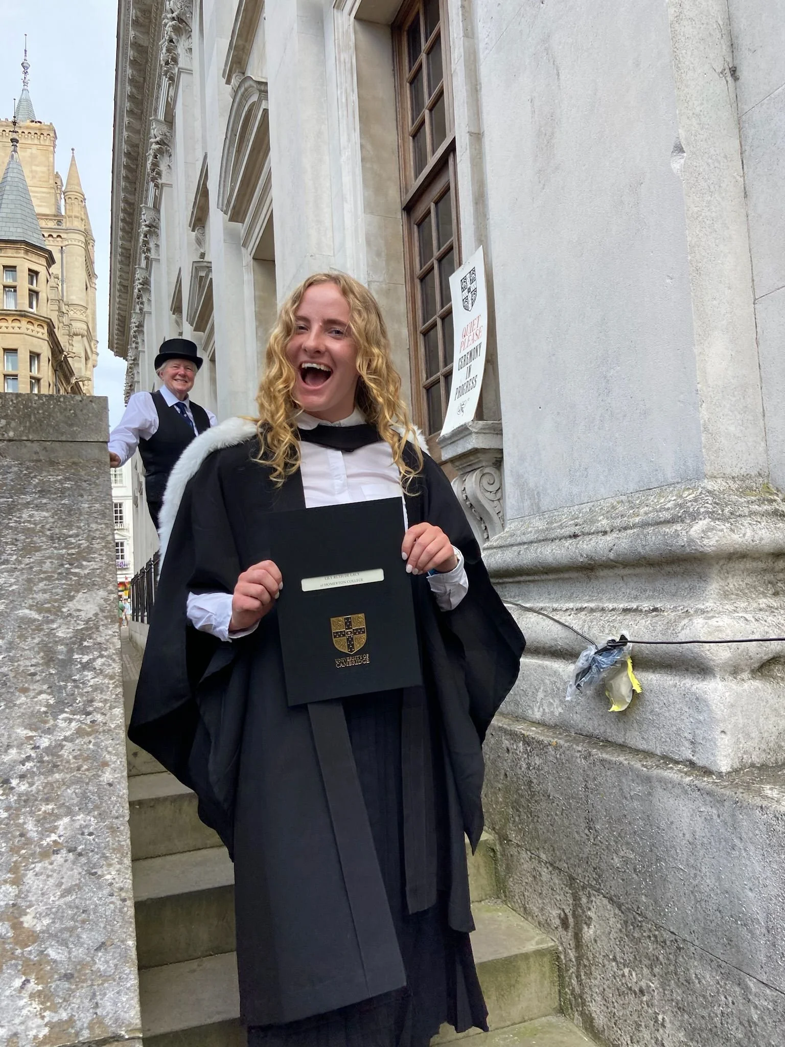 Young women in graduation robes and caps standing outside a historic building, one holding a diploma, smiling and celebrating.