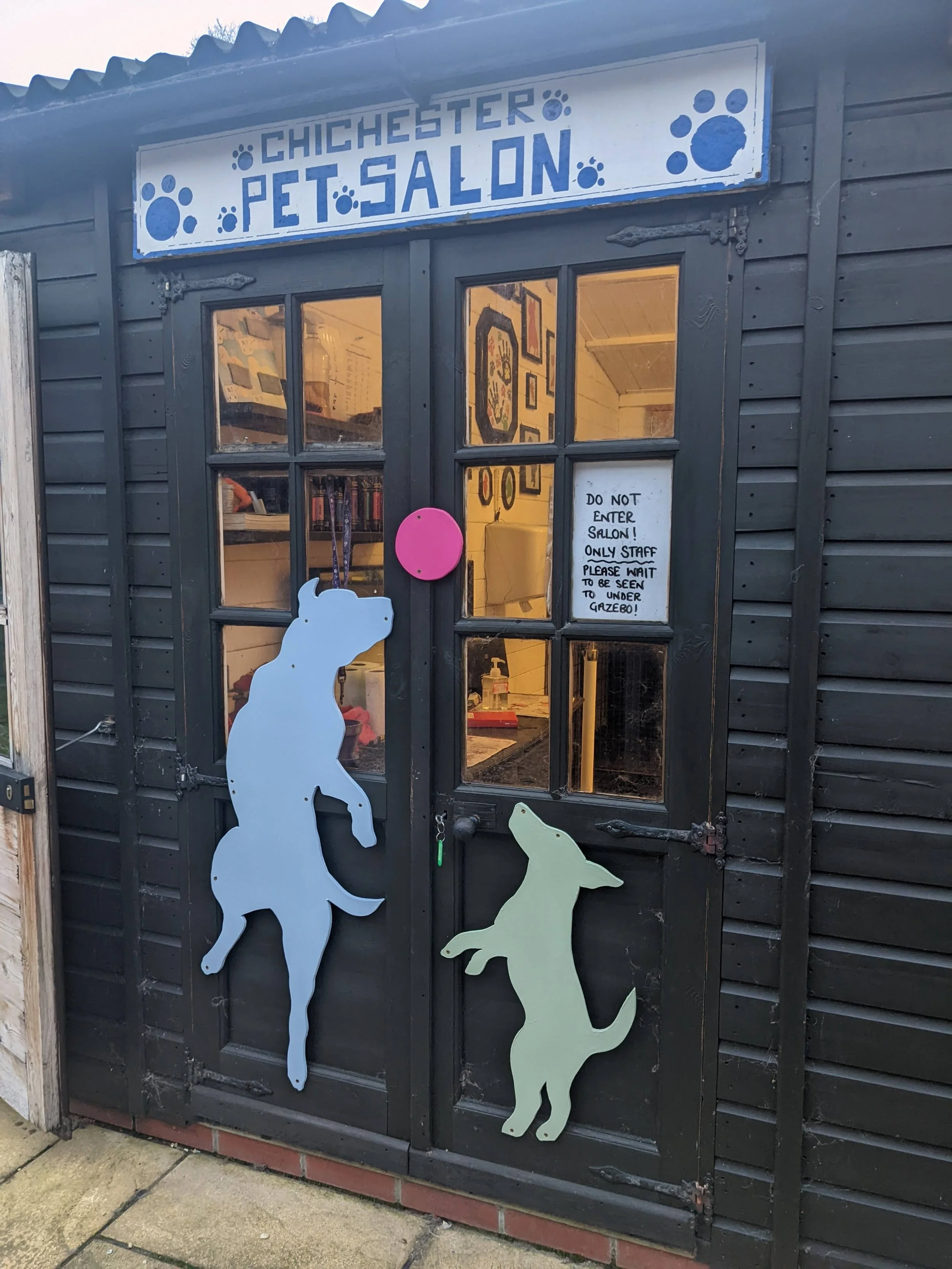 Exterior of a black wooden pet salon with a sign reading 'Chichester Pet Salon' decorated with blue paw prints. The door has animal cutouts, one of a large dog or wolf and a smaller dog. A pink circular sign and a handwritten note on the door say, 'D