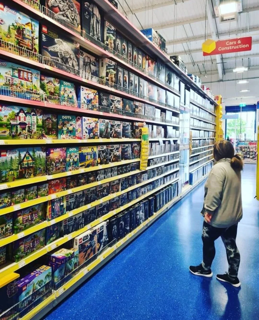 A woman shopping for Lego sets in a store aisle with shelves filled with various Lego building kits.
