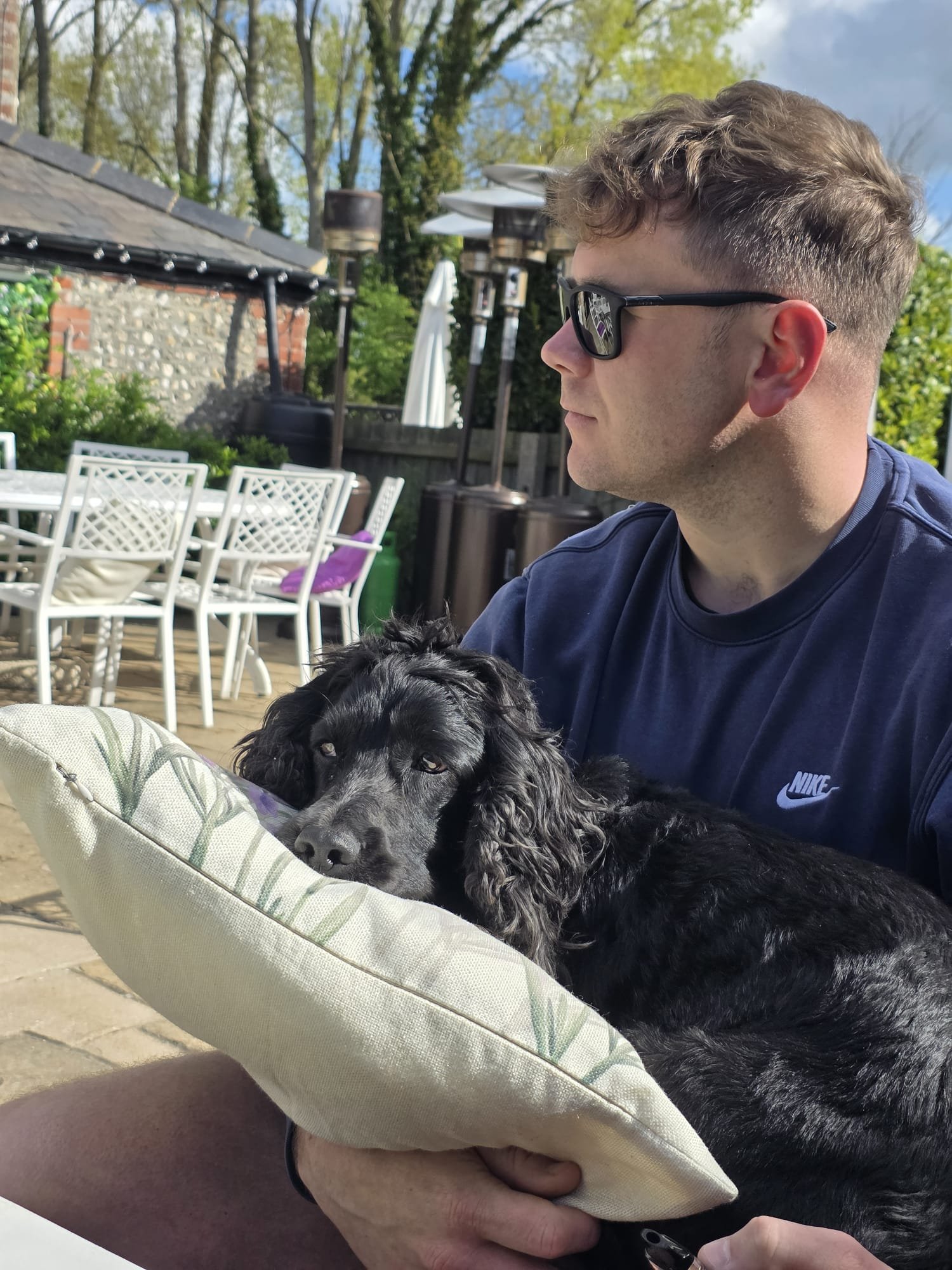 Man wearing sunglasses and a Nike shirt sitting outdoors with a black dog resting on a cushioned seat.