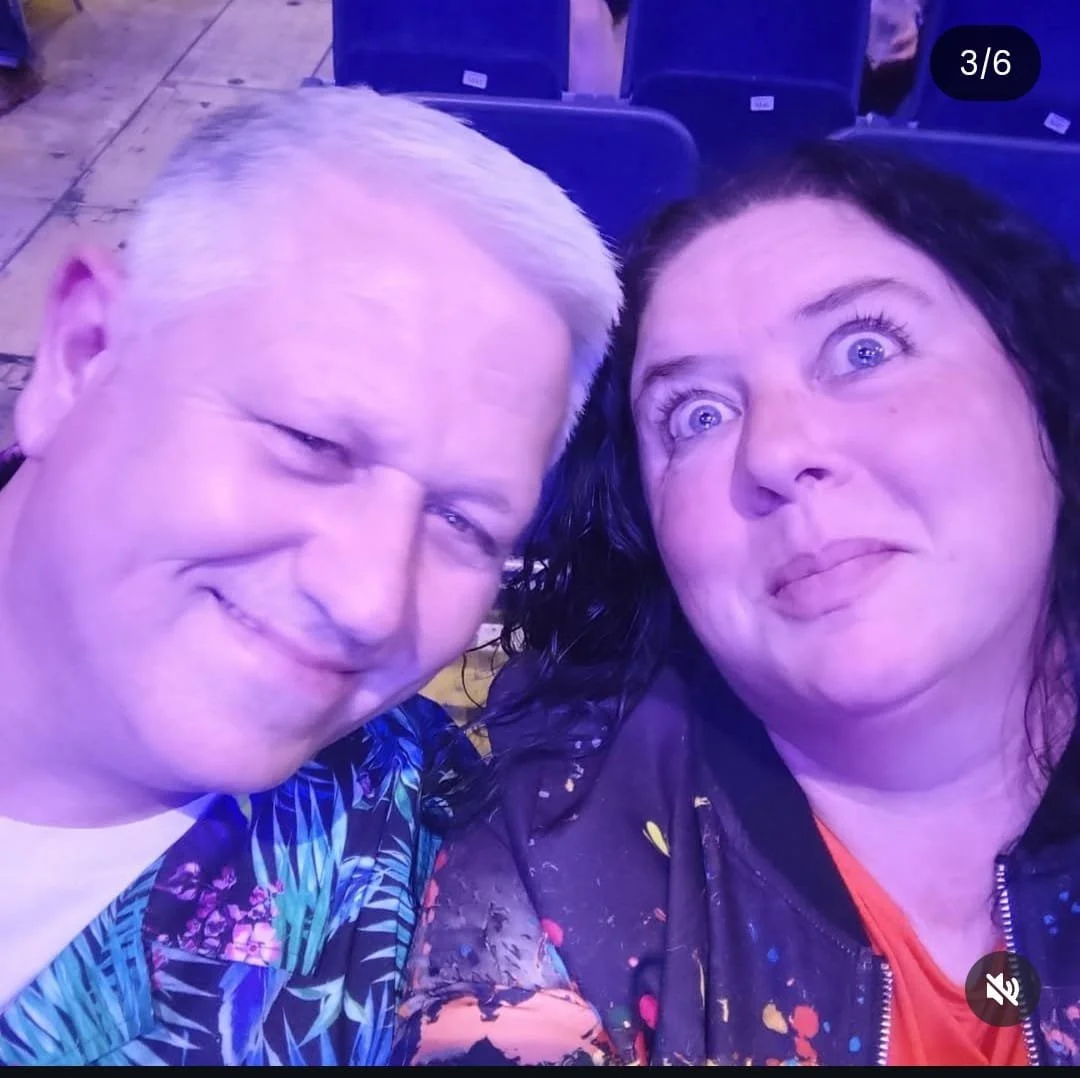 Couple taking a selfie at an indoor event, with purple lighting and empty blue chairs in the background.