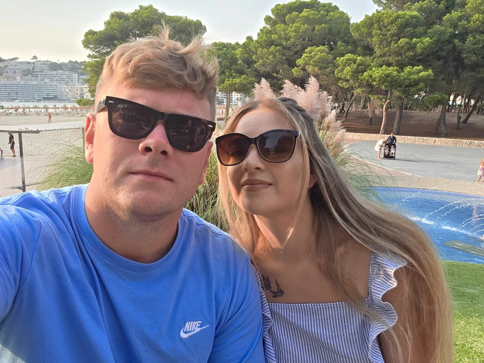 A couple wearing sunglasses taking a selfie outdoors at a park near the beach, with trees, a fountain, and people in the background.
