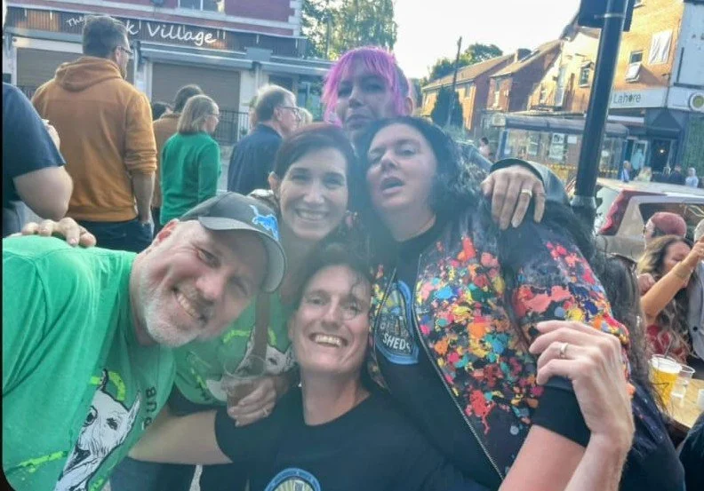 Group of five friends outdoors, smiling and hugging at a lively event, with booths and other people in the background.