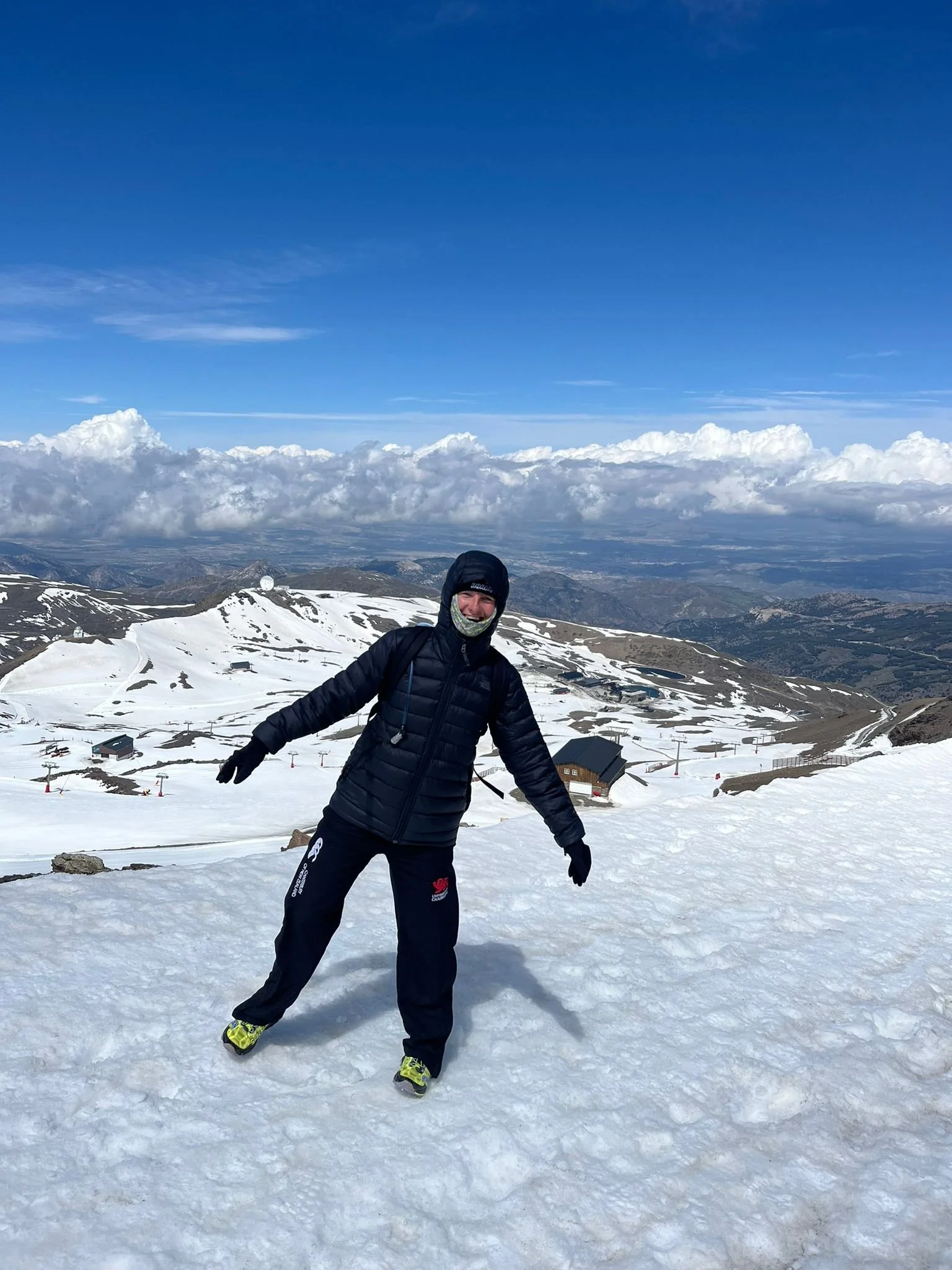 A person in winter clothing standing on snow-covered ground on a mountain with a panoramic view of snow patches, buildings, and winding paths under a partly cloudy blue sky.