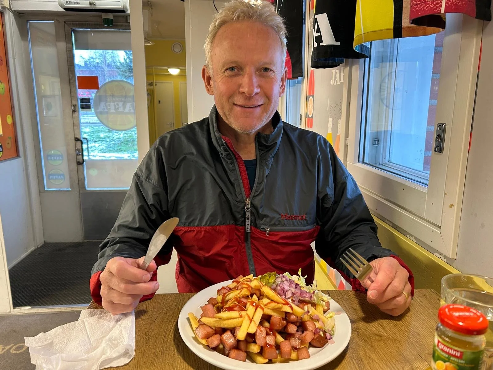 A man with gray hair smiling while sitting at a table inside a restaurant, holding a knife in his left hand and a fork in his right hand, with a plate of hot dogs topped with fries and chopped onions in front of him.