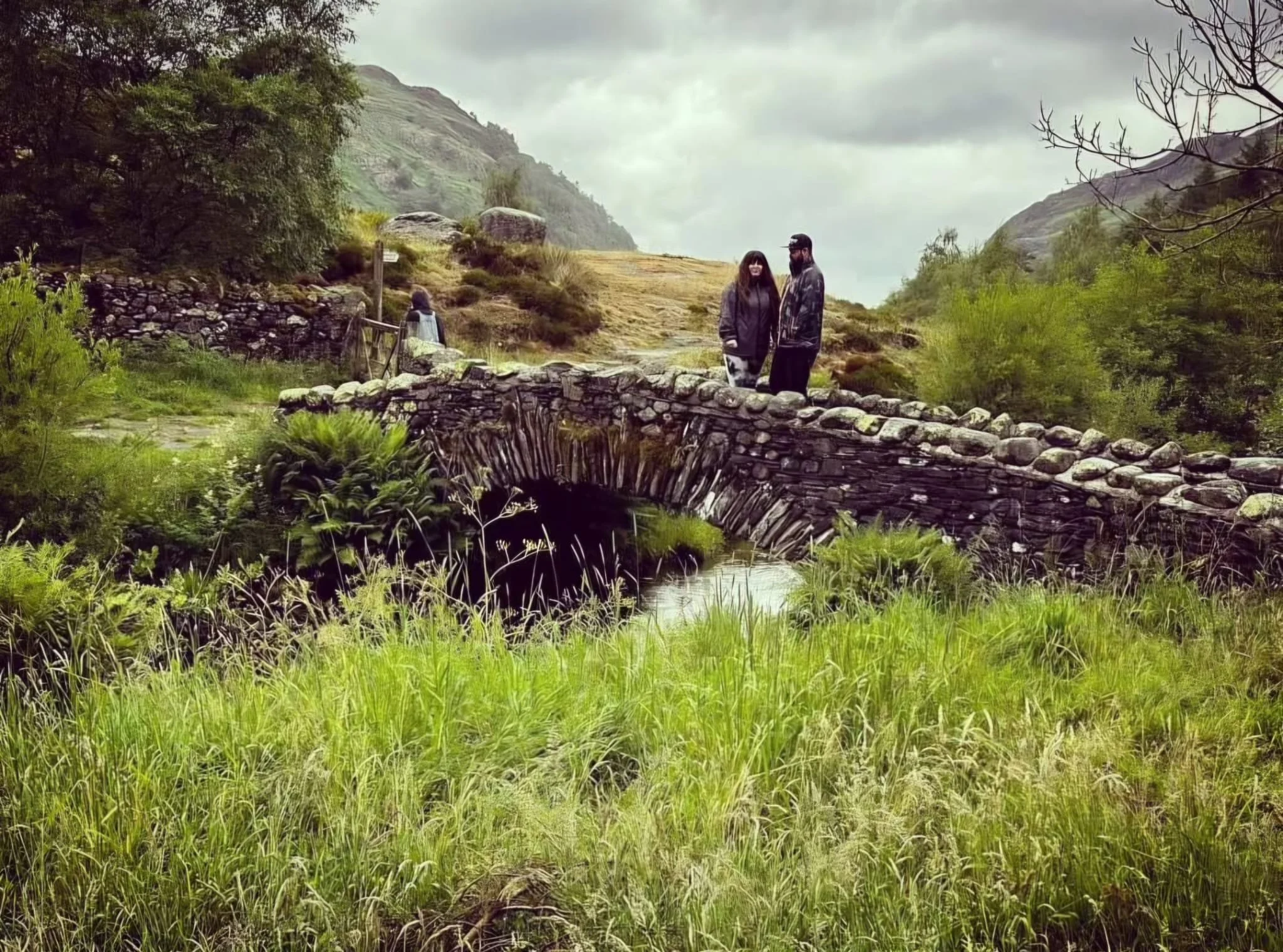 Two people standing on a stone bridge over a small creek in a lush, green, mountain landscape with cloudy sky at the lake district.