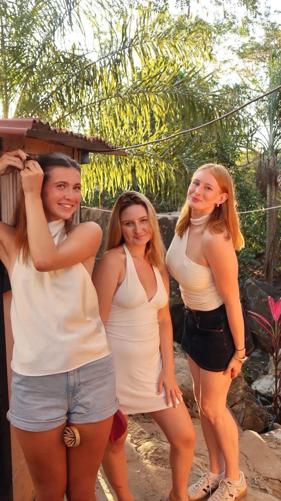 Three young women standing outdoors in a lush, tropical setting with palm trees and plants, smiling and posing for the photo during sunset.