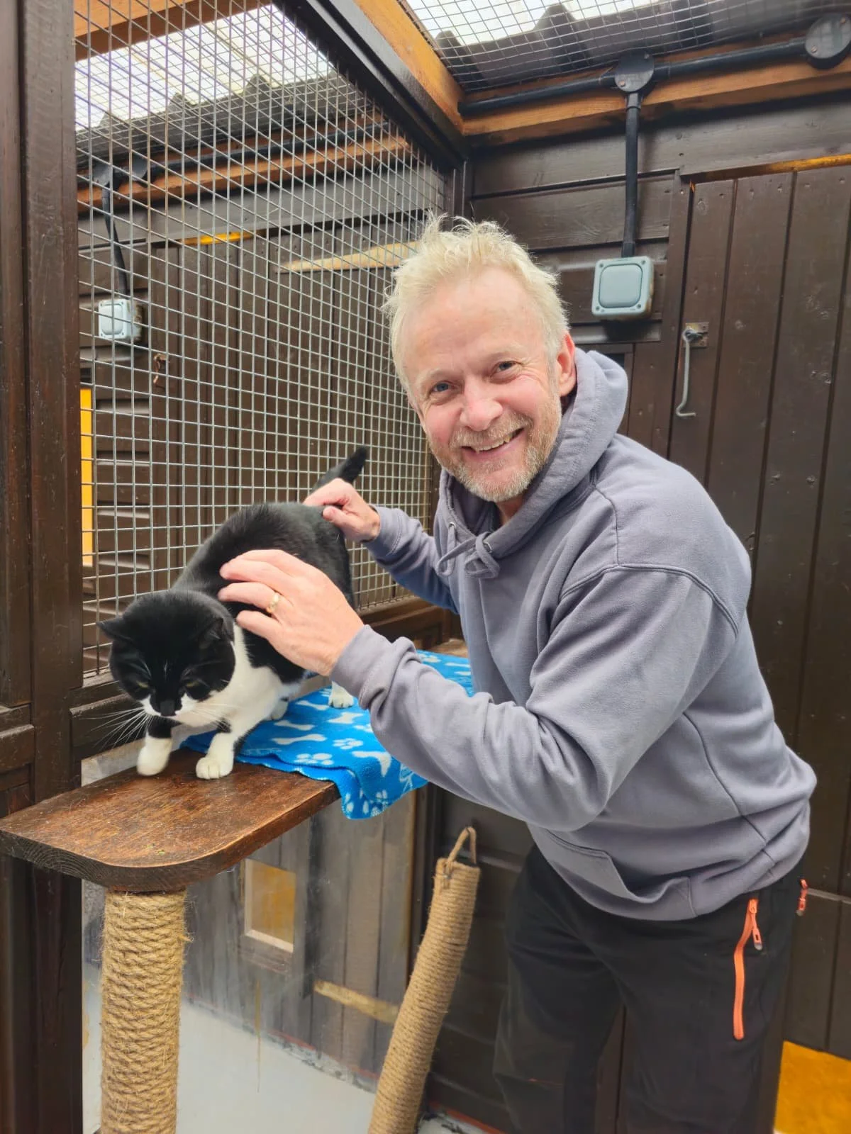 A man with blonde hair and a gray hoodie smiling while petting a black and white cat inside a wooden and wire enclosure.