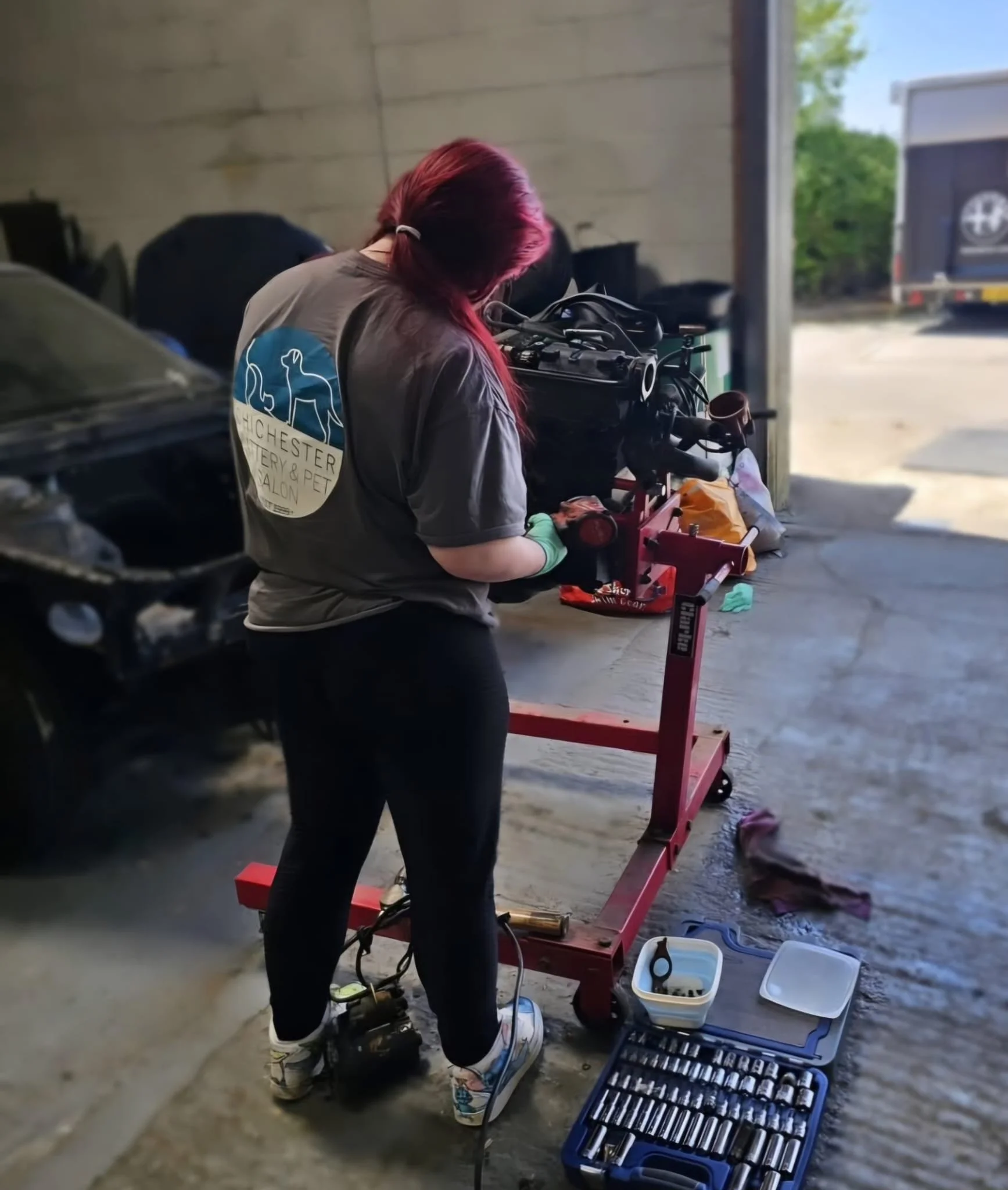 A person with red hair working on a car engine inside a garage, with tools and equipment on the ground nearby.