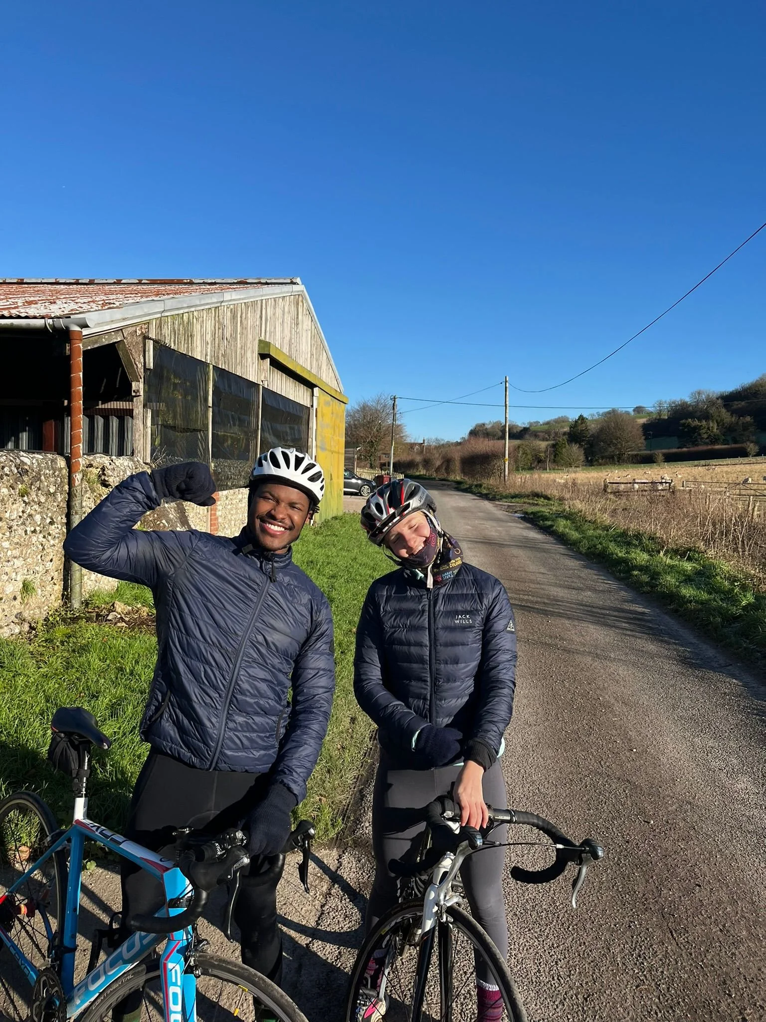 Two women with cycling helmets and jackets smiling and posing with their bikes on a rural road, with one flexing her arm and the other leaning her head. Clear blue sky and countryside scenery in the background.