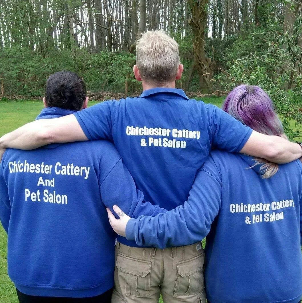 Three people wearing blue shirts with white text that reads 'Chichester Cattery & Pet Salon,' standing outdoors with their arms around each other, facing away from the camera in a grassy area with trees in the background.