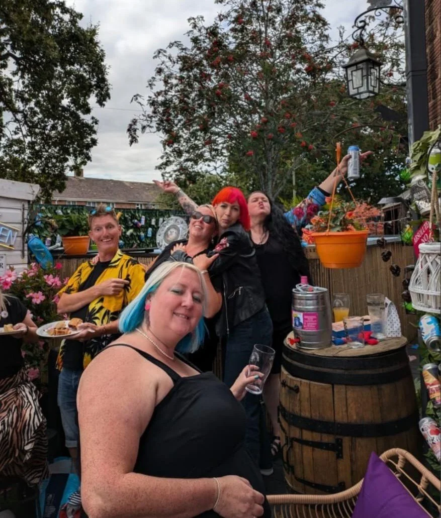 A group of friends celebrating outdoors on a backyard patio, with trees and a cloudy sky in the background. They are smiling, holding drinks, and posing for the photo. Decorations, plants, and outdoor furniture are visible.