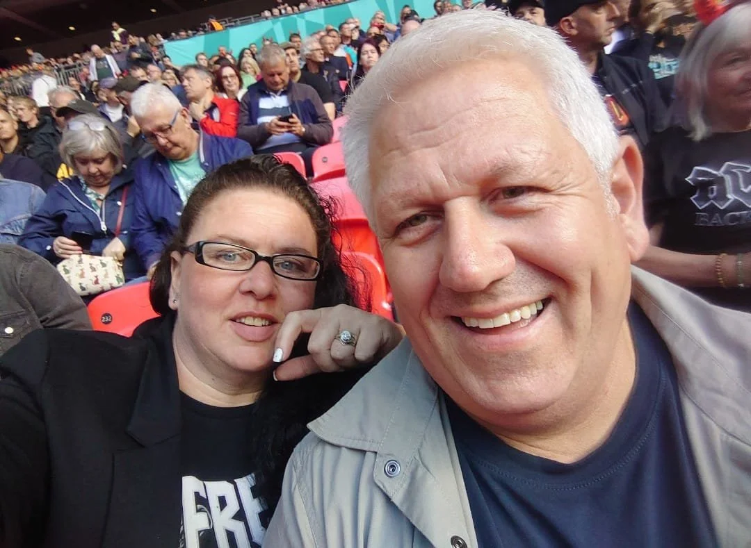 A group of people seated at an event, with two individuals in the foreground taking a selfie. The woman has dark hair, glasses, and a black jacket, while the man has white hair and is smiling broadly.