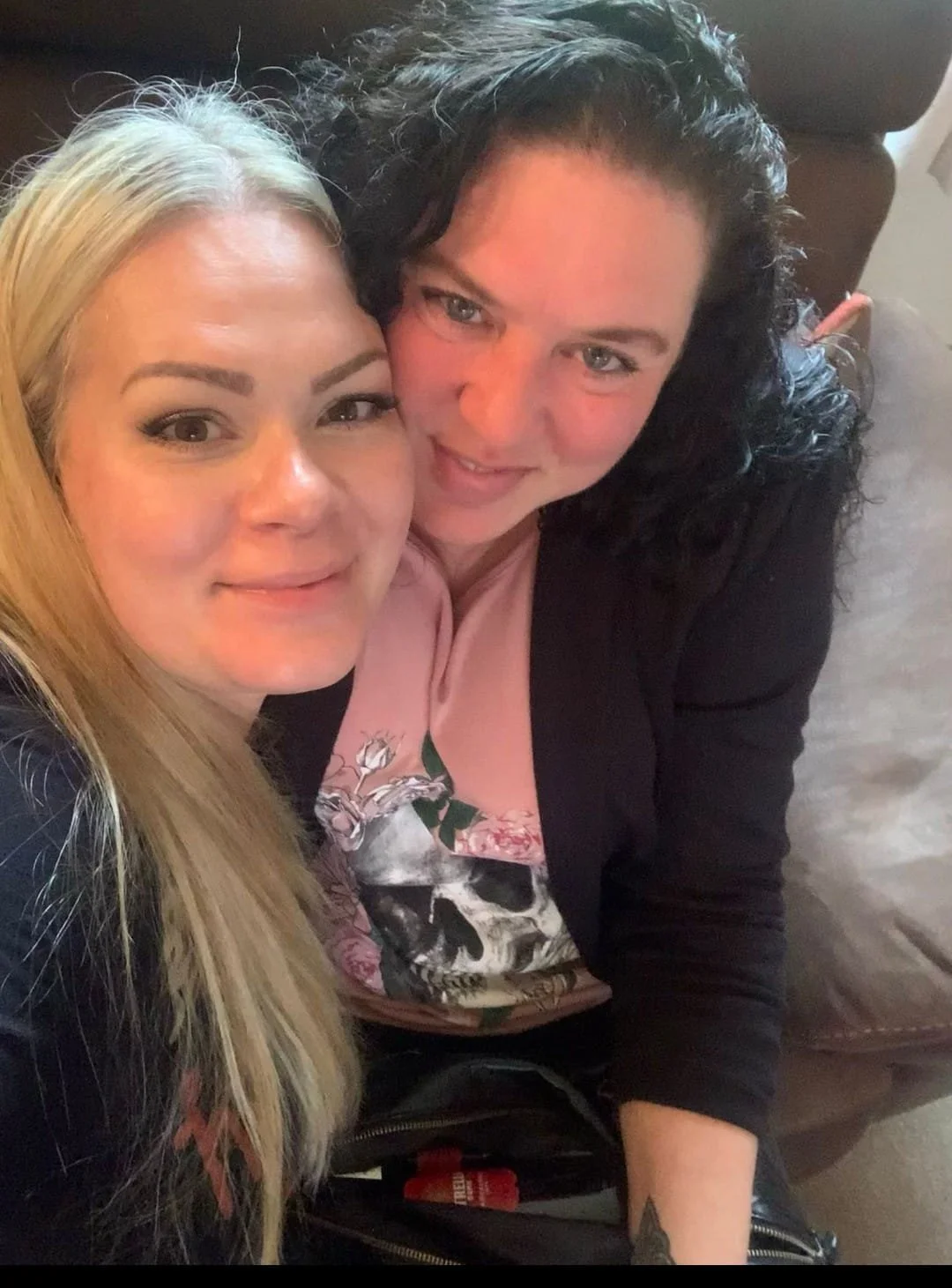 Two women sitting on a brown couch, taking a selfie together indoors.