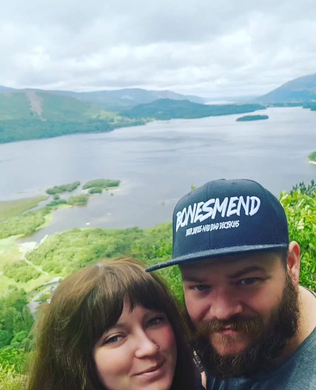 A couple taking a selfie overlooking a scenic landscape with a large body of water, green hills, and mountains in the background. The woman has brown hair and the man is wearing a black cap with white text.