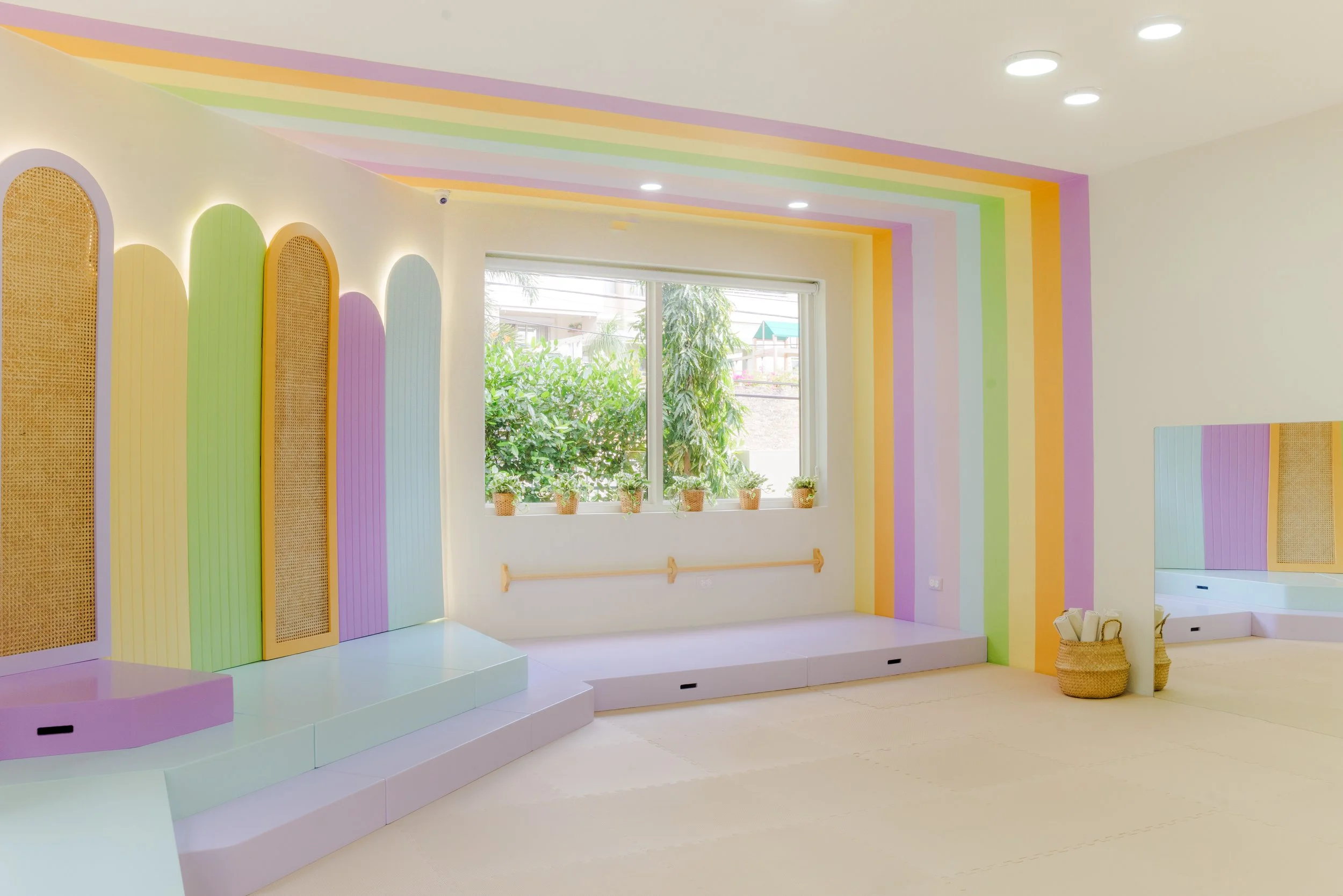 Sala decorada con paredes y techo en colores pastel, con un gran ventanal con plantas en la ventana y piso de color beis, diseñada para niños.