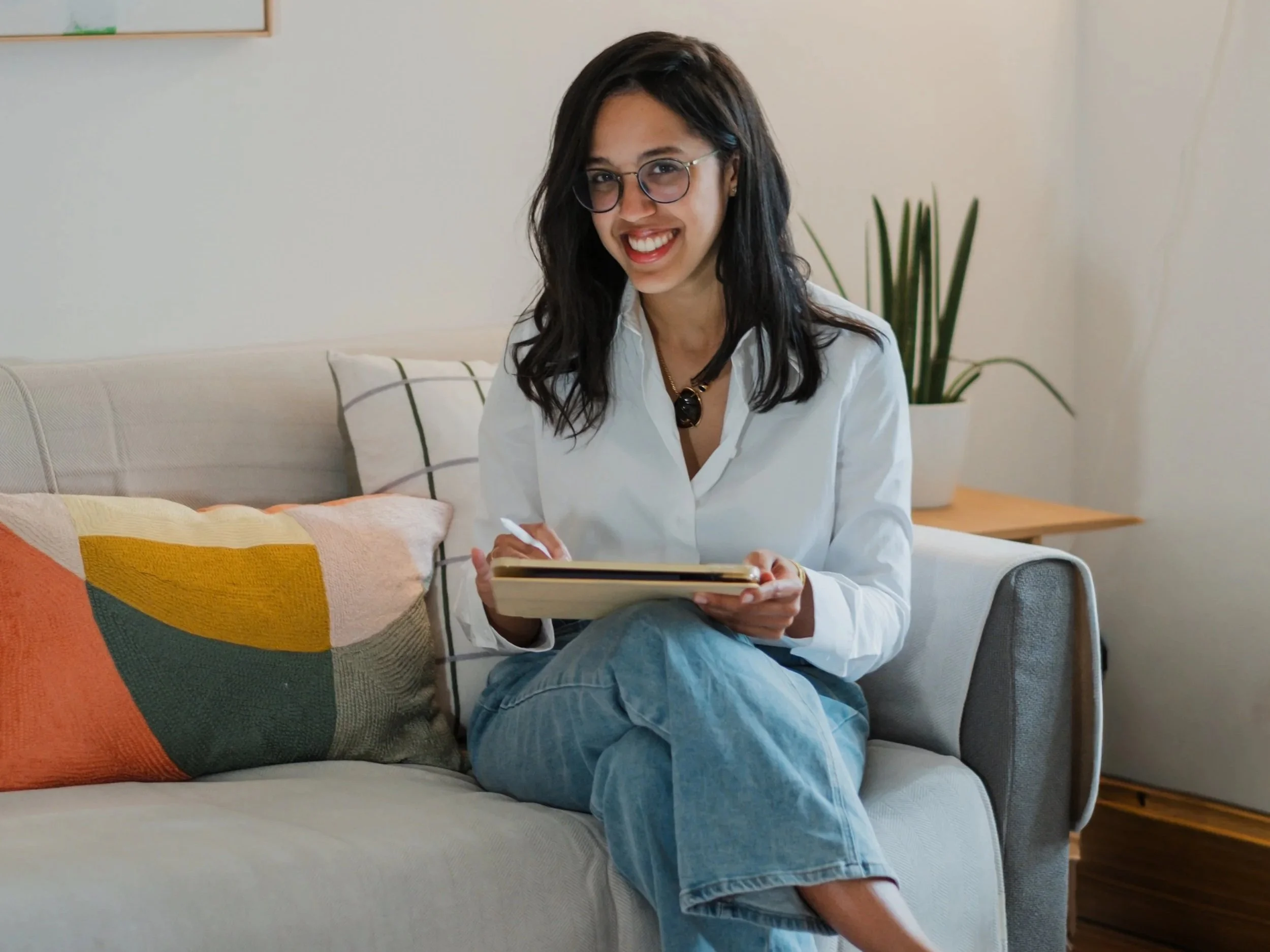 Mujer joven con gafas, camiseta blanca y pantalones vaqueros sentada en un sofá, sonriendo y escribiendo en un cuaderno.