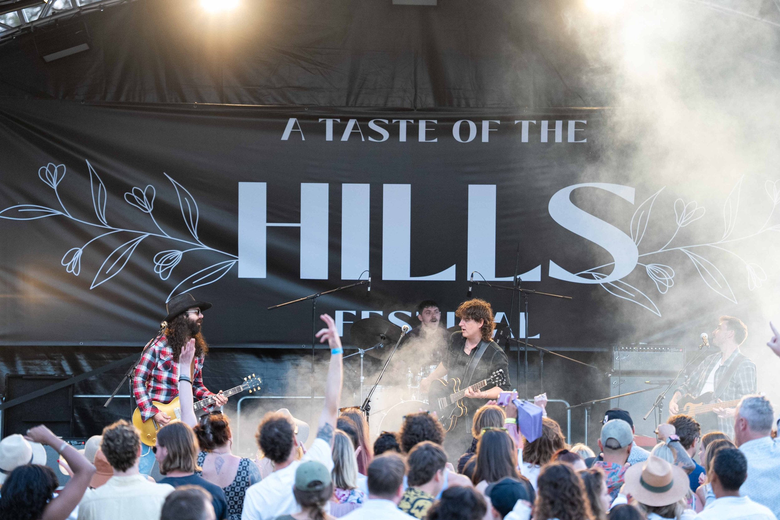 A live outdoor music performance at a festival named A Taste Of The Hills Festival, with a band playing on stage and a large banner in the background displaying the festival's name, surrounded by an audience.