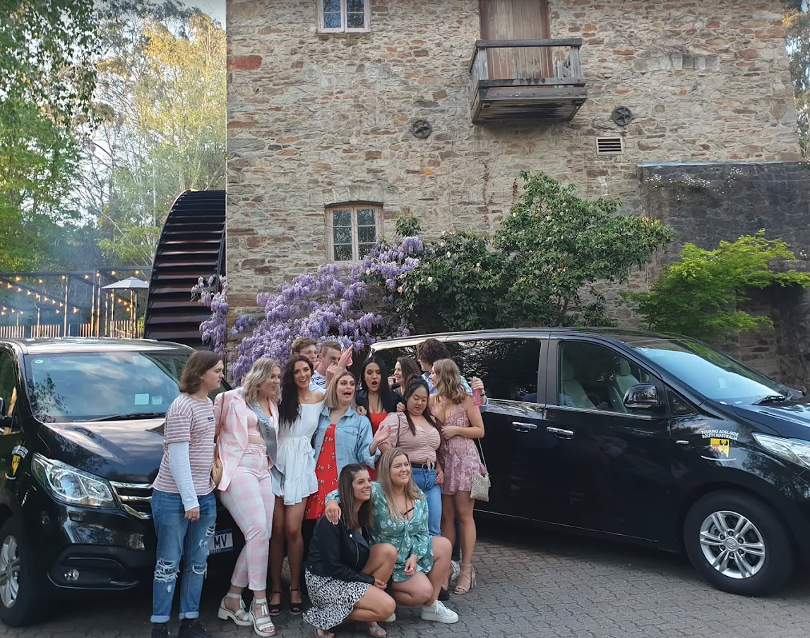A group of people posing for a photo in front of parked cars and a stone building with purple flowers and greenery.