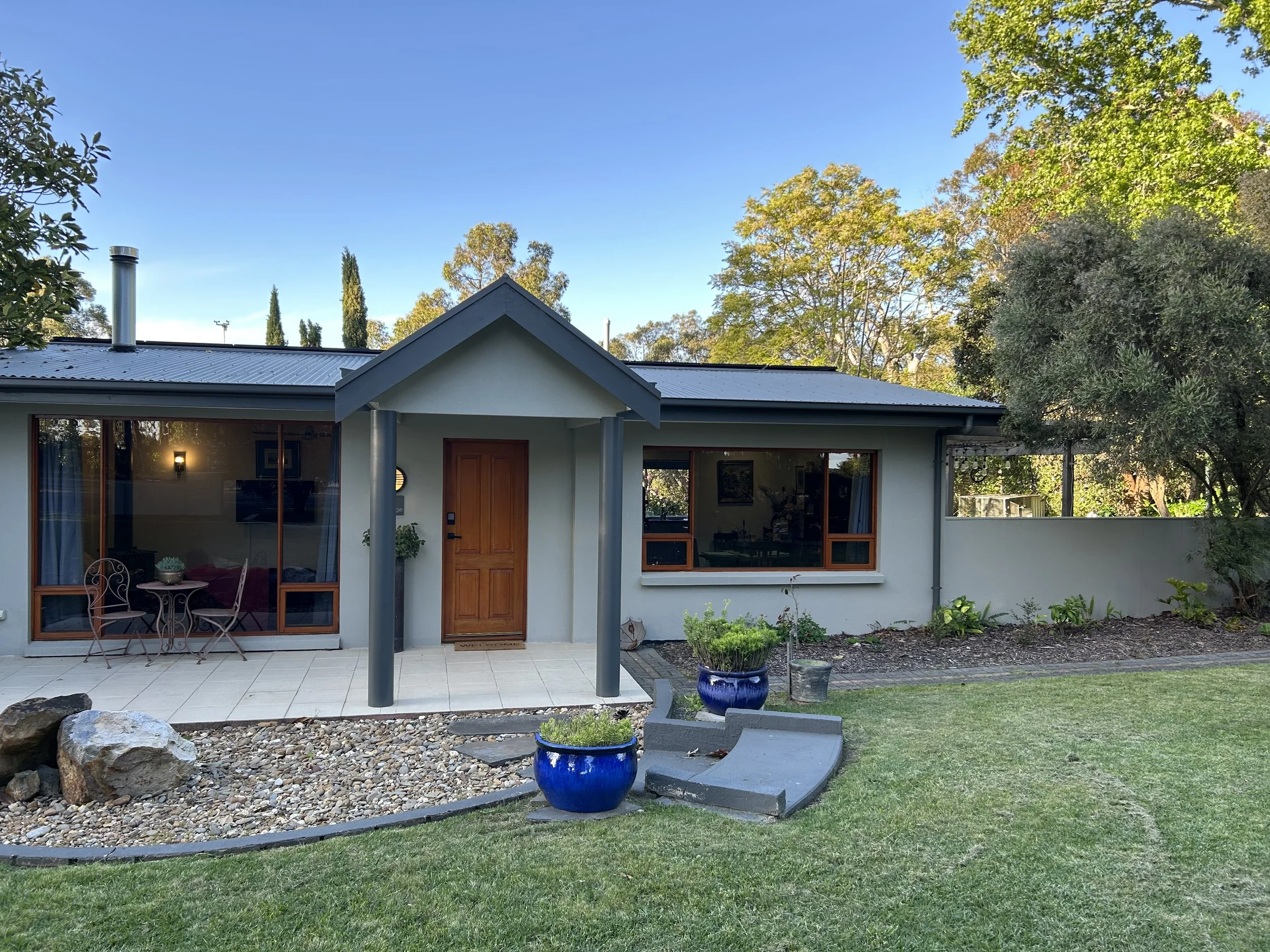 Front view of a single-story house with a porch, featuring a wooden door, large windows, and a surrounding garden with green grass, potted plants, rocks, and trees in the background under a clear blue sky.