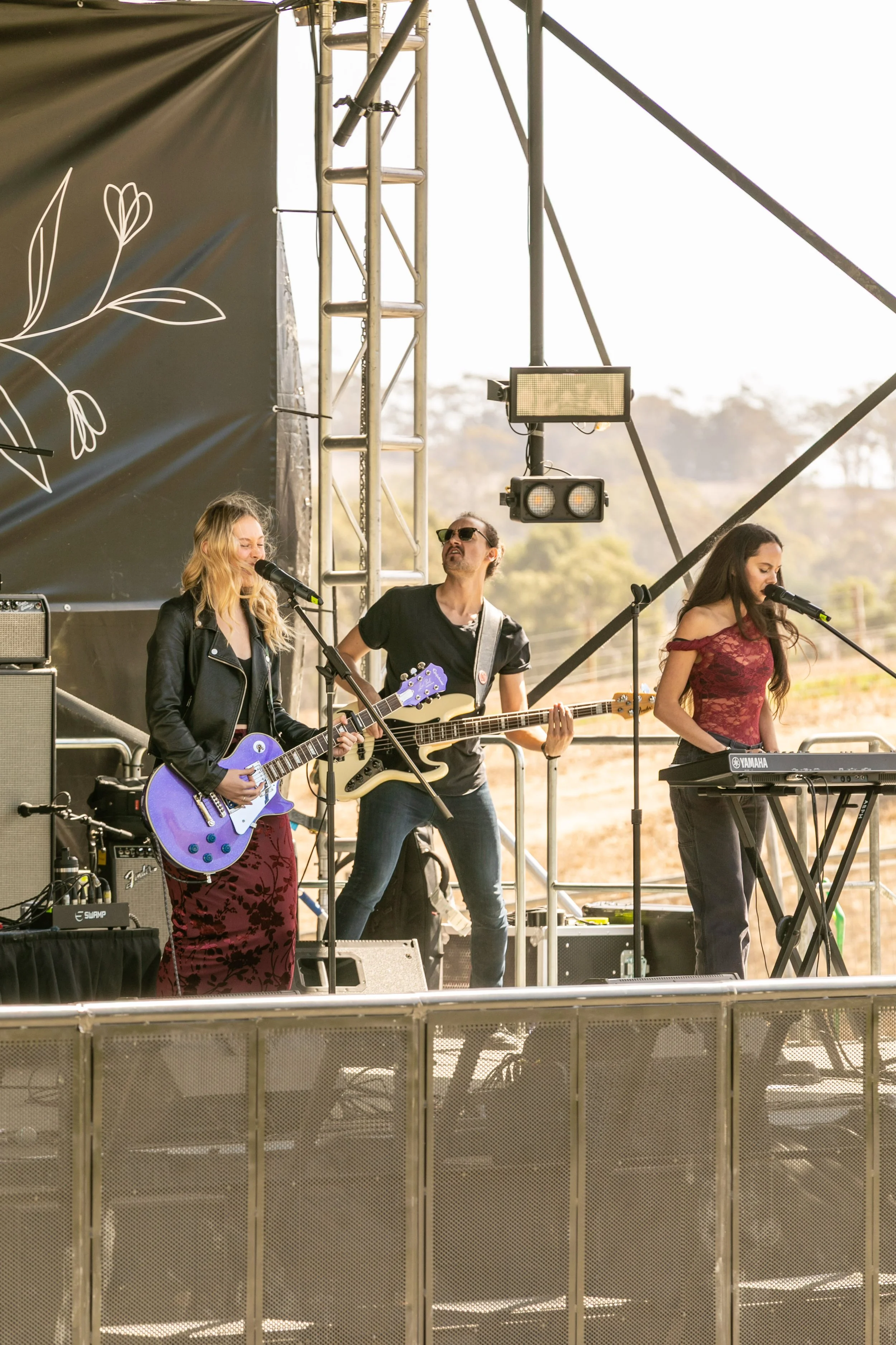 Three musicians perform on an outdoor stage. The woman on the left is playing a purple electric guitar and singing into a microphone, the man in the middle is playing a bass guitar, and the woman on the right is singing into a microphone while playing a keyboard.