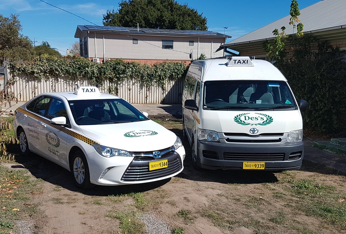 A white Toyota taxi car and a white Toyota van taxi parked on a dirt lot in front of a fence with bushes and a house in the background.
