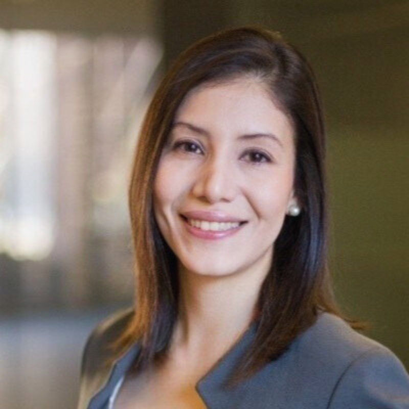 Portrait of a young woman with shoulder-length dark curly hair, wearing a white top with black stripes, smiling softly with blue blurred background.