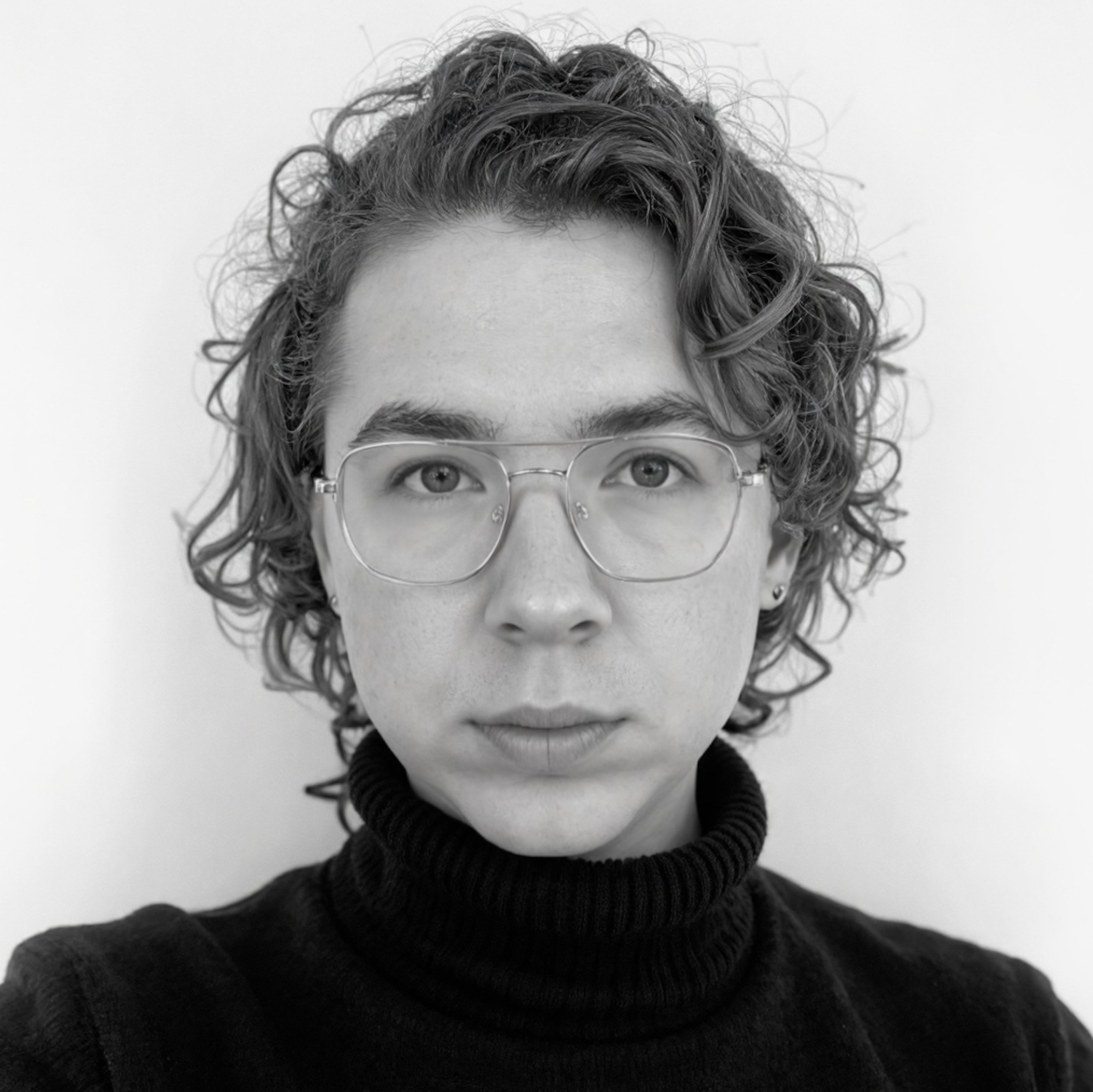 Close-up photo of a young person with curly hair, glasses, and a gentle smile, wearing a white shirt and holding their chin with one hand.