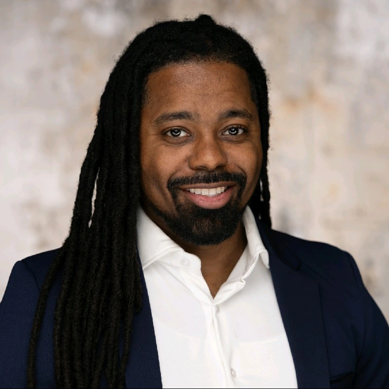 A man with long dreadlocks, glasses, and a beard, dressed in a purple suit, sitting on a chair in an indoor setting with large windows and a brown floor.