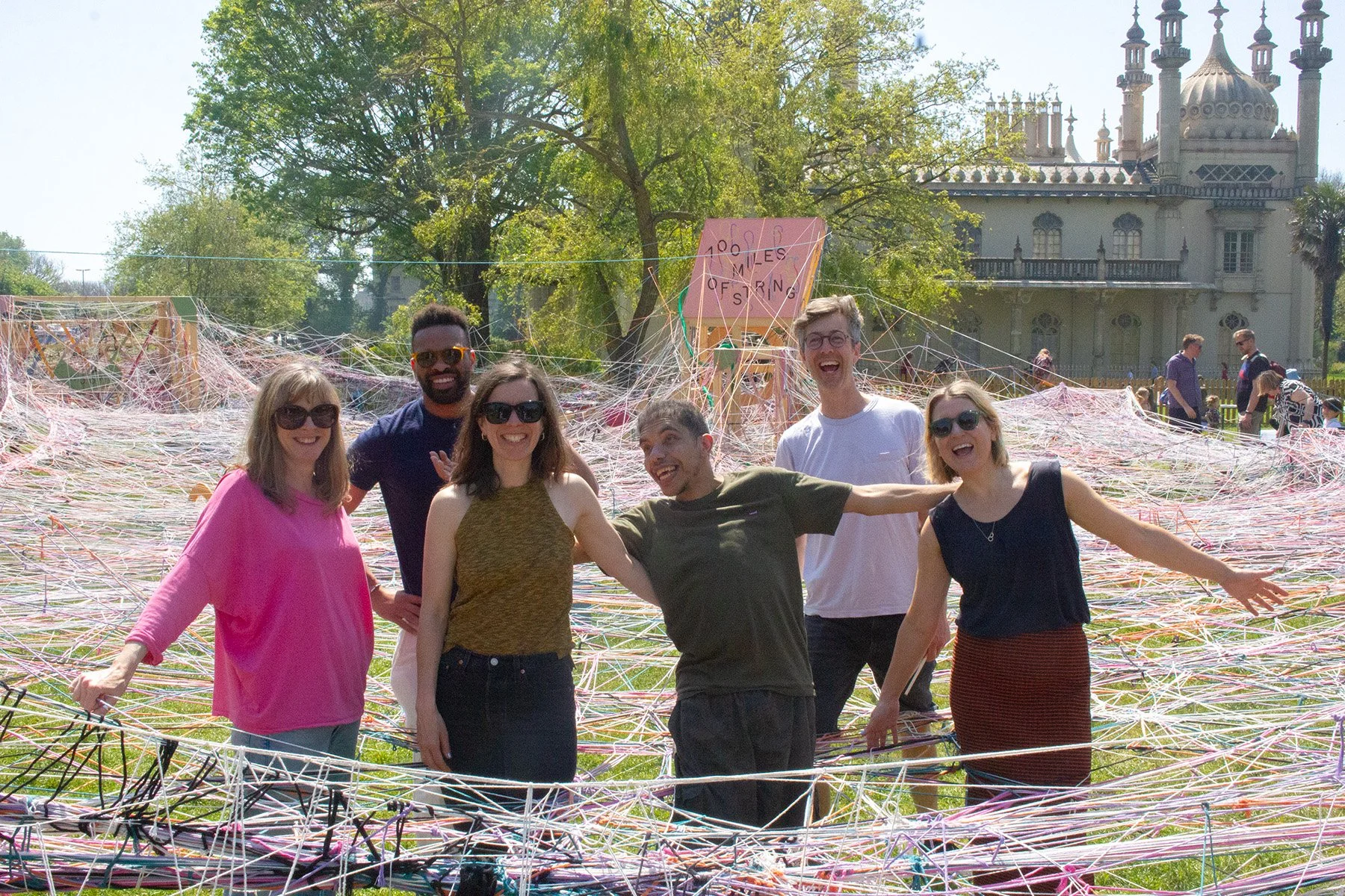 A group of six friends smiling and posing in front of an art installation made of colorful yarn and string, with a historic building and trees in the background.