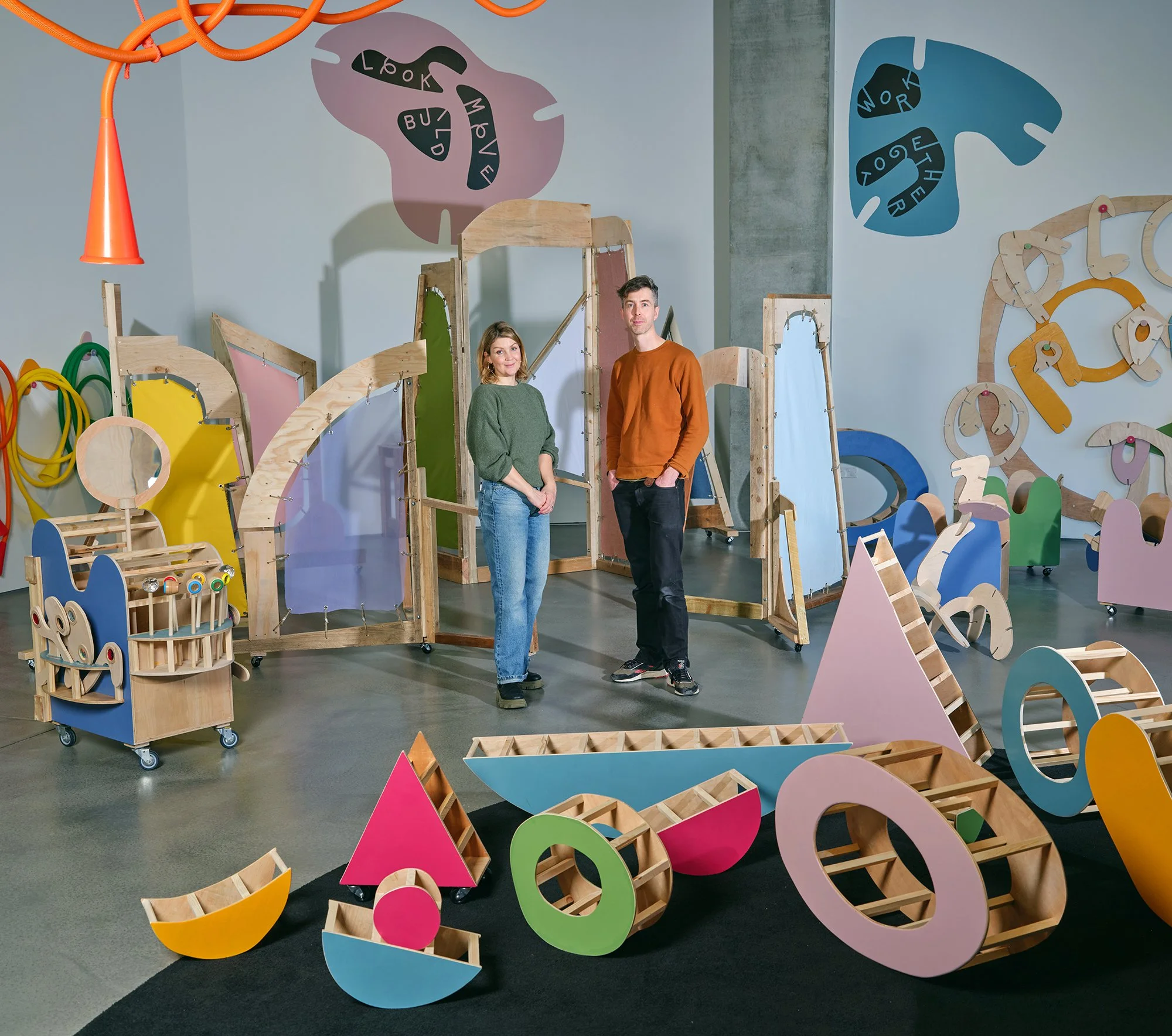 Two people standing in an indoor play area with colorful, abstract wooden and foam structures for children to play with.