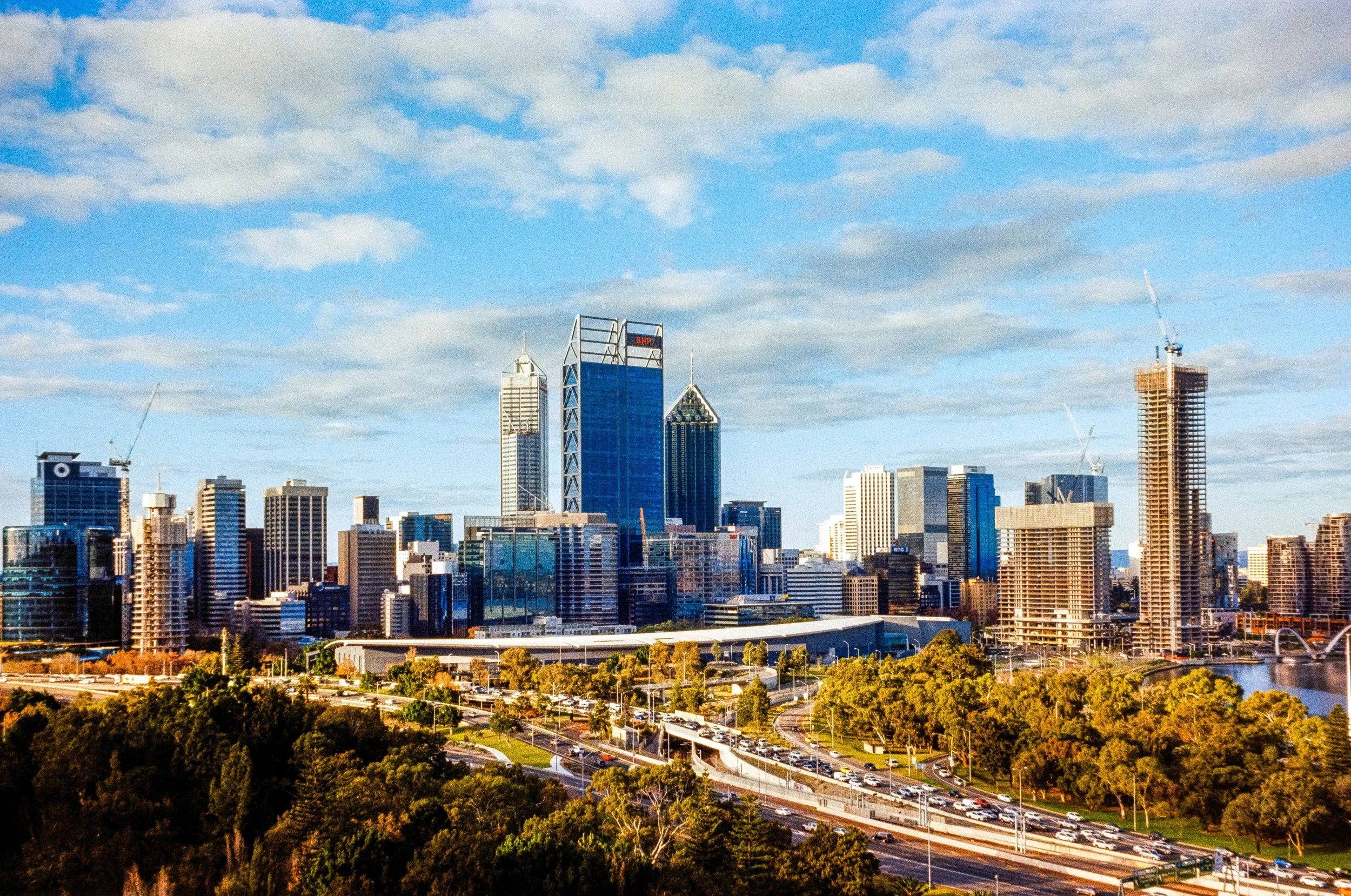 A city skyline with modern skyscrapers set against a blue sky with scattered clouds, with greenery, roads, and traffic in the foreground.