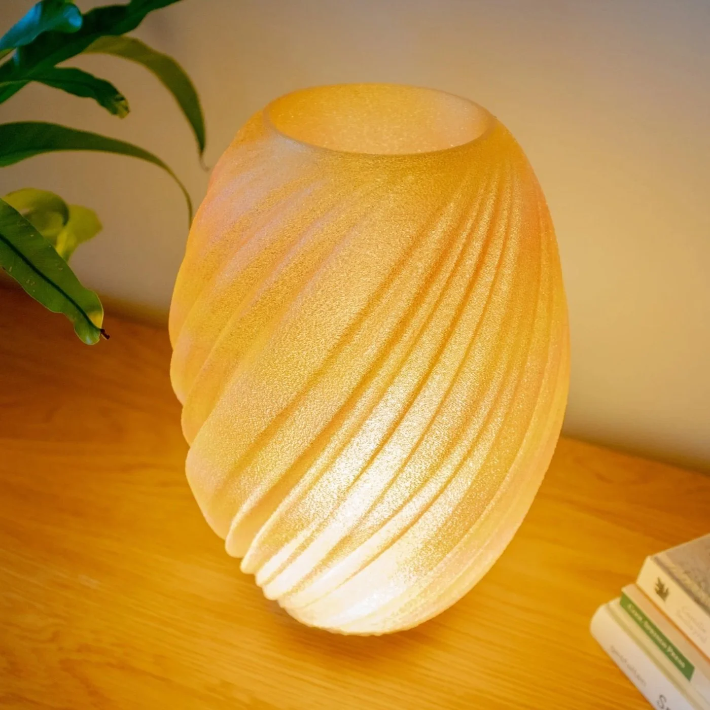 A glowing, oval yellow 3d printed lamp with a textured surface, sitting on a modern white tripod base on a wooden table, with a potted plant in a white textured pot in the background.