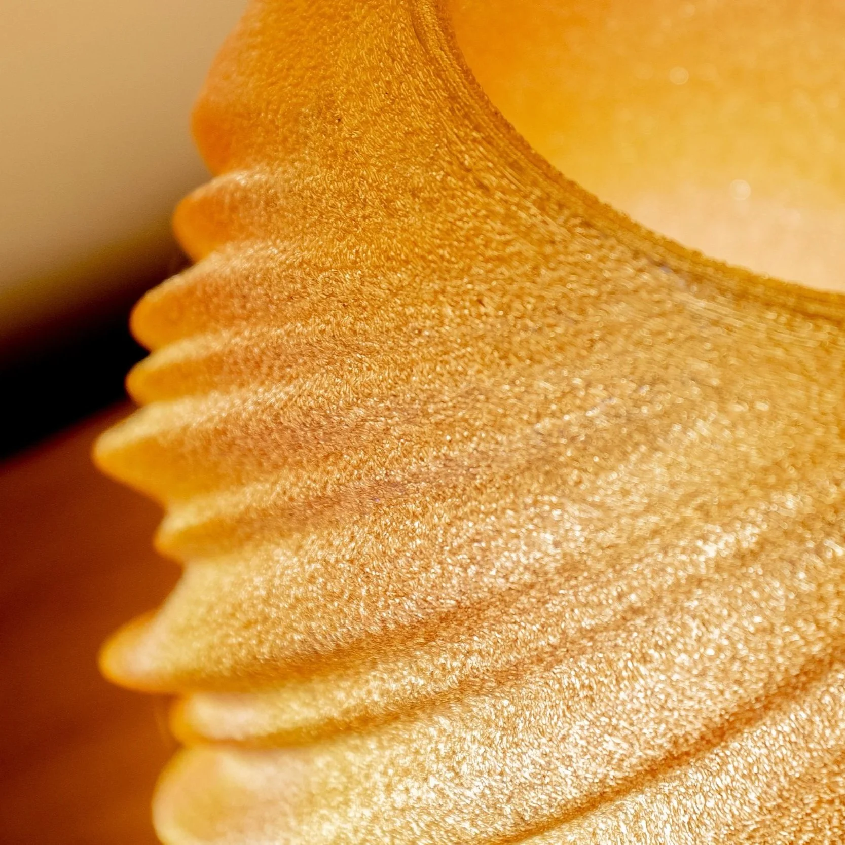 Close-up of a ridged, yellow 3d printed lamp with a textured surface, sitting on a modern white tripod base on a wooden table, with a potted plant in a white textured pot in the background.