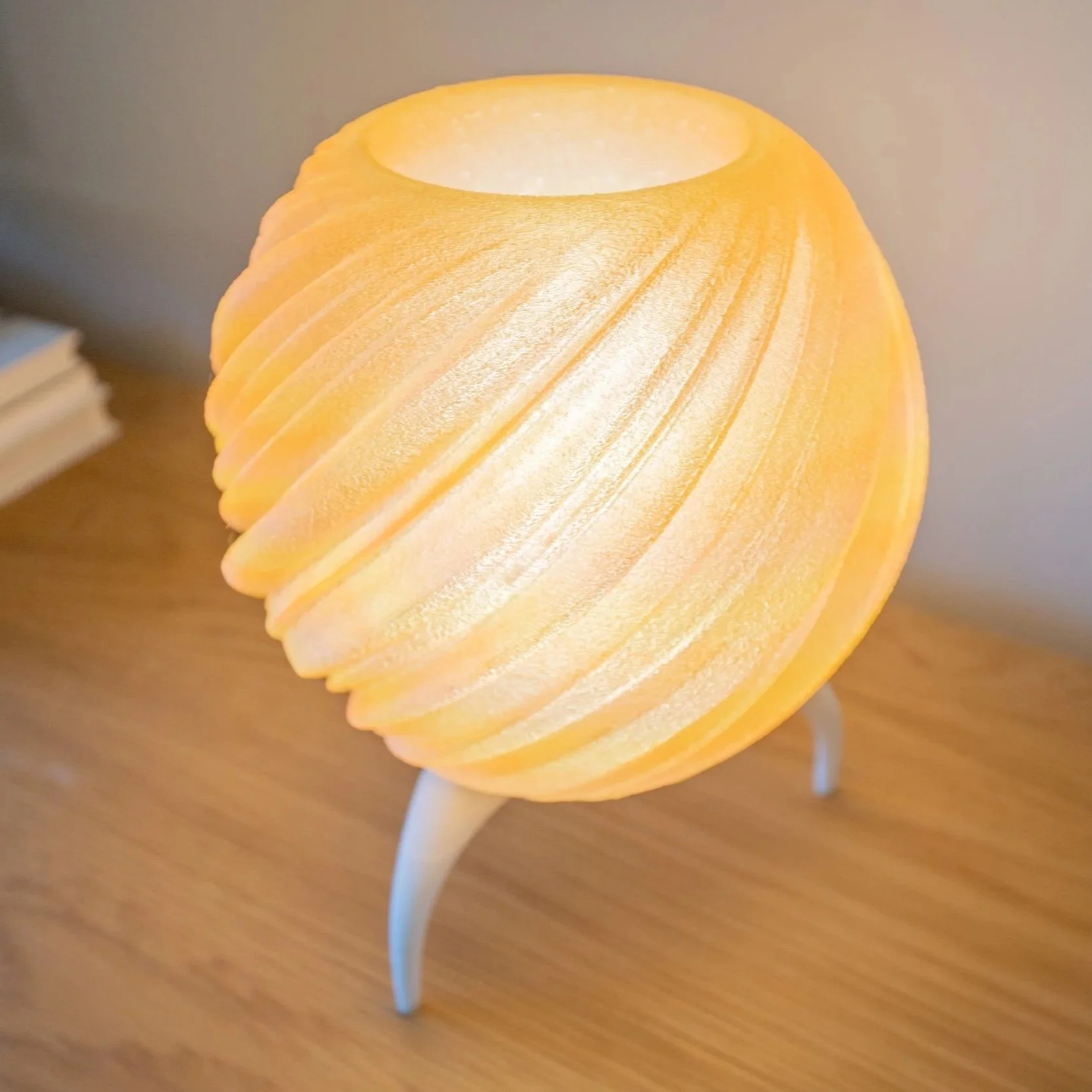 A glowing, spherical yellow 3d printed lamp with a textured surface, sitting on a modern white tripod base on a wooden table, with a potted plant in a white textured pot in the background.